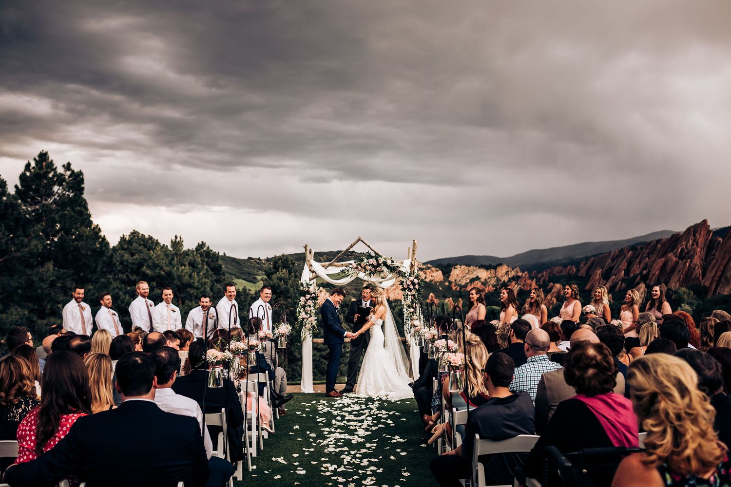 Dramatic mountain backdrop frames an outdoor wedding ceremony with decorated white arch and seated guests.