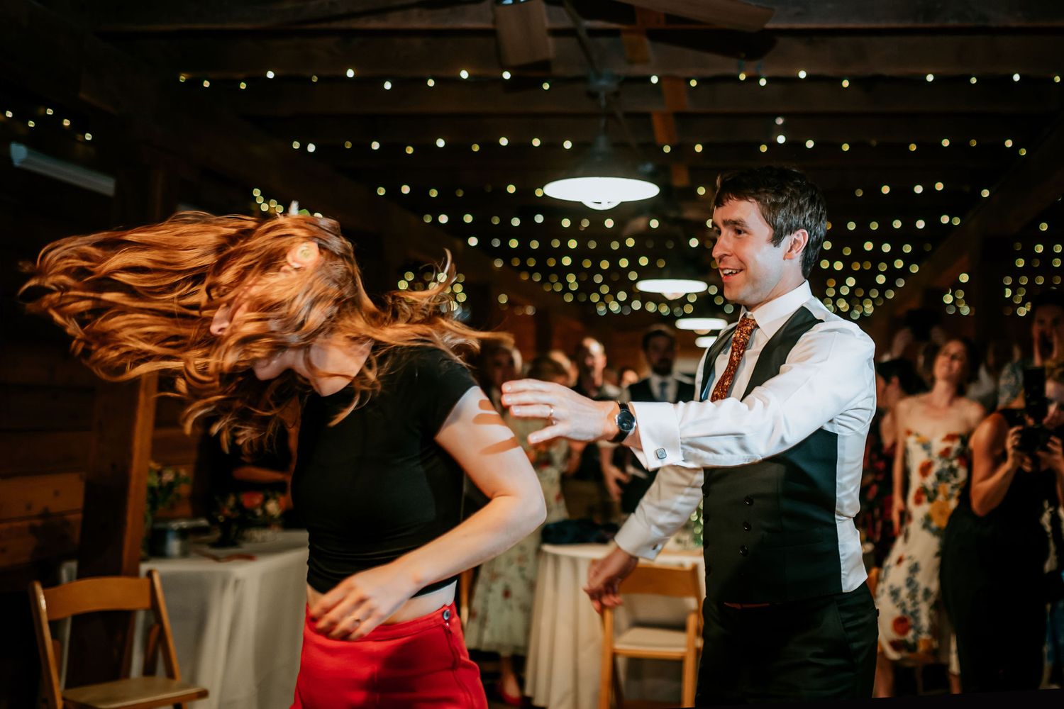 Couple dances energetically at wedding reception with string lights overhead.