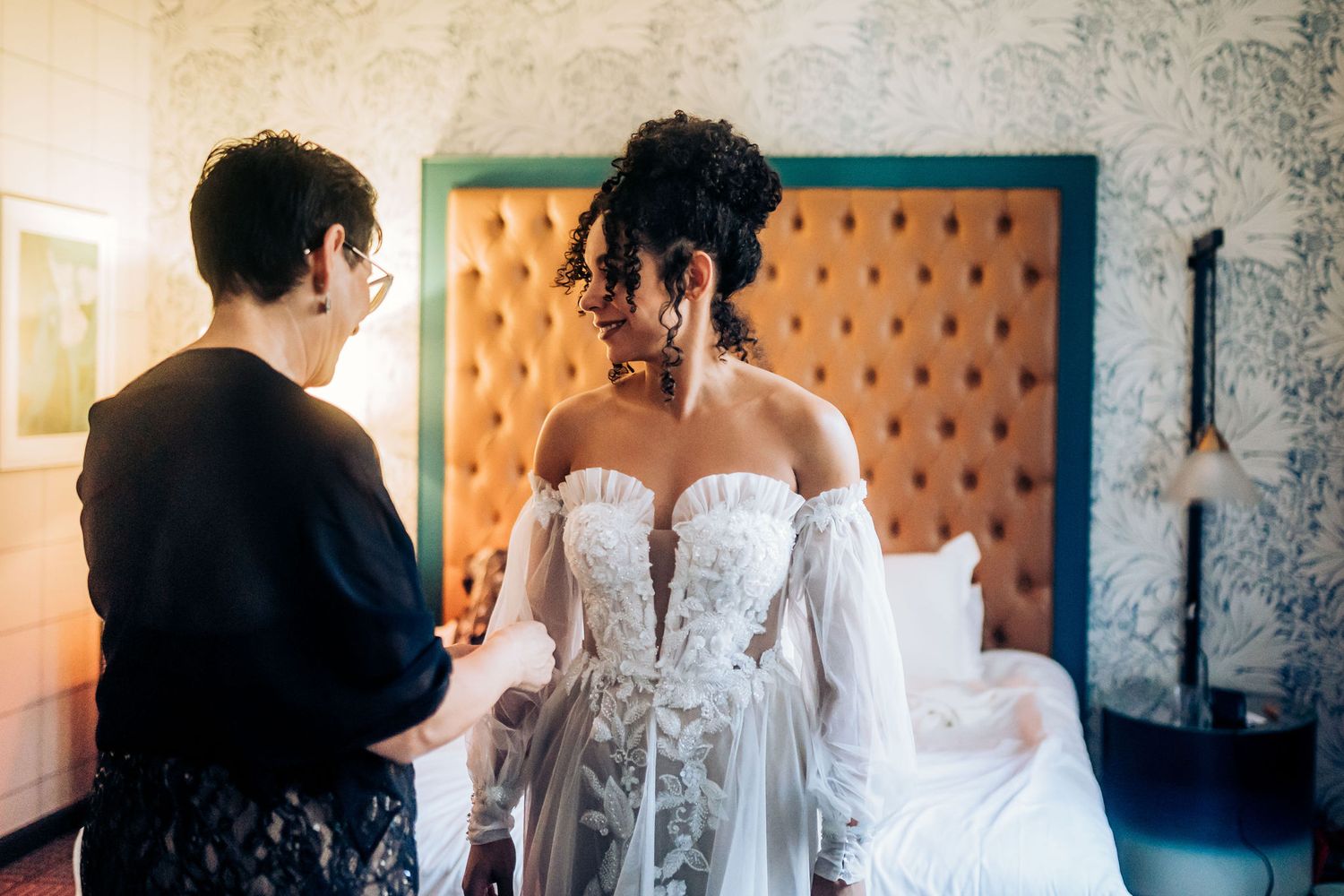 A detailed white wedding dress being fitted in a room with vintage-style orange tufted headboard.