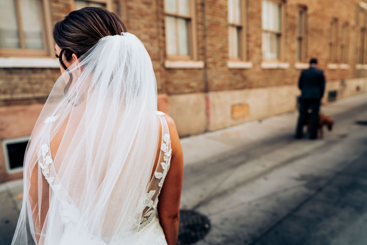 A flowing white veil and lace wedding dress from behind on a city street.