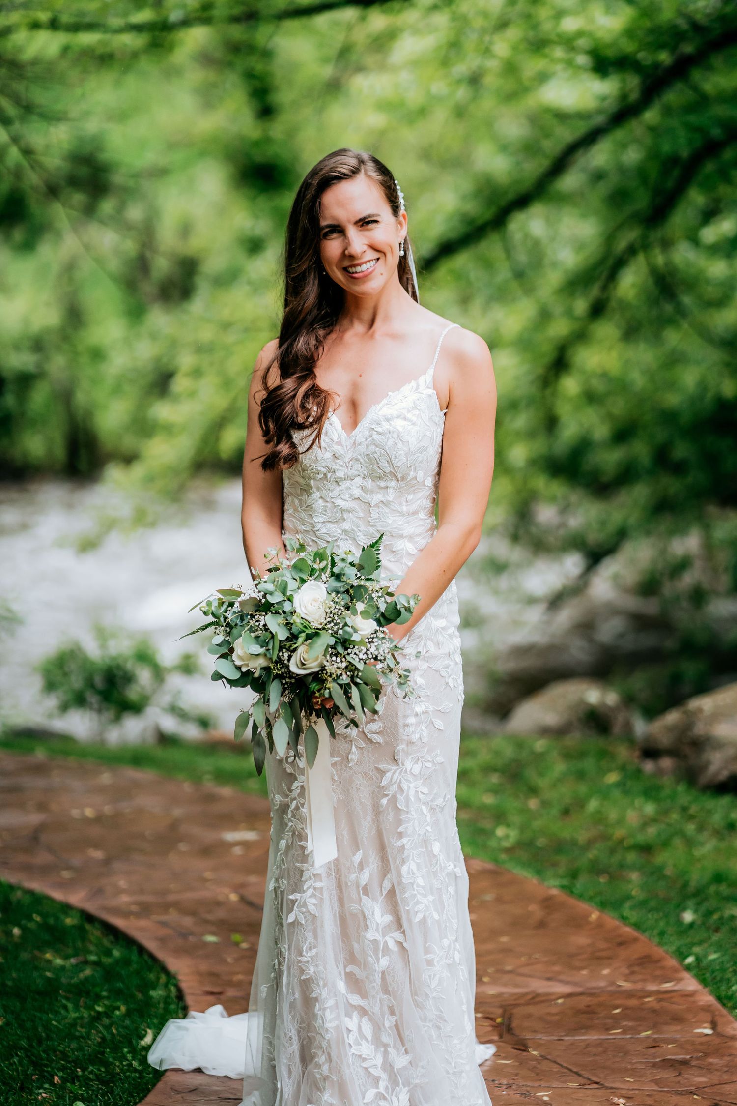 Bride in lace wedding gown holds elegant white and green bouquet on stone path by river.