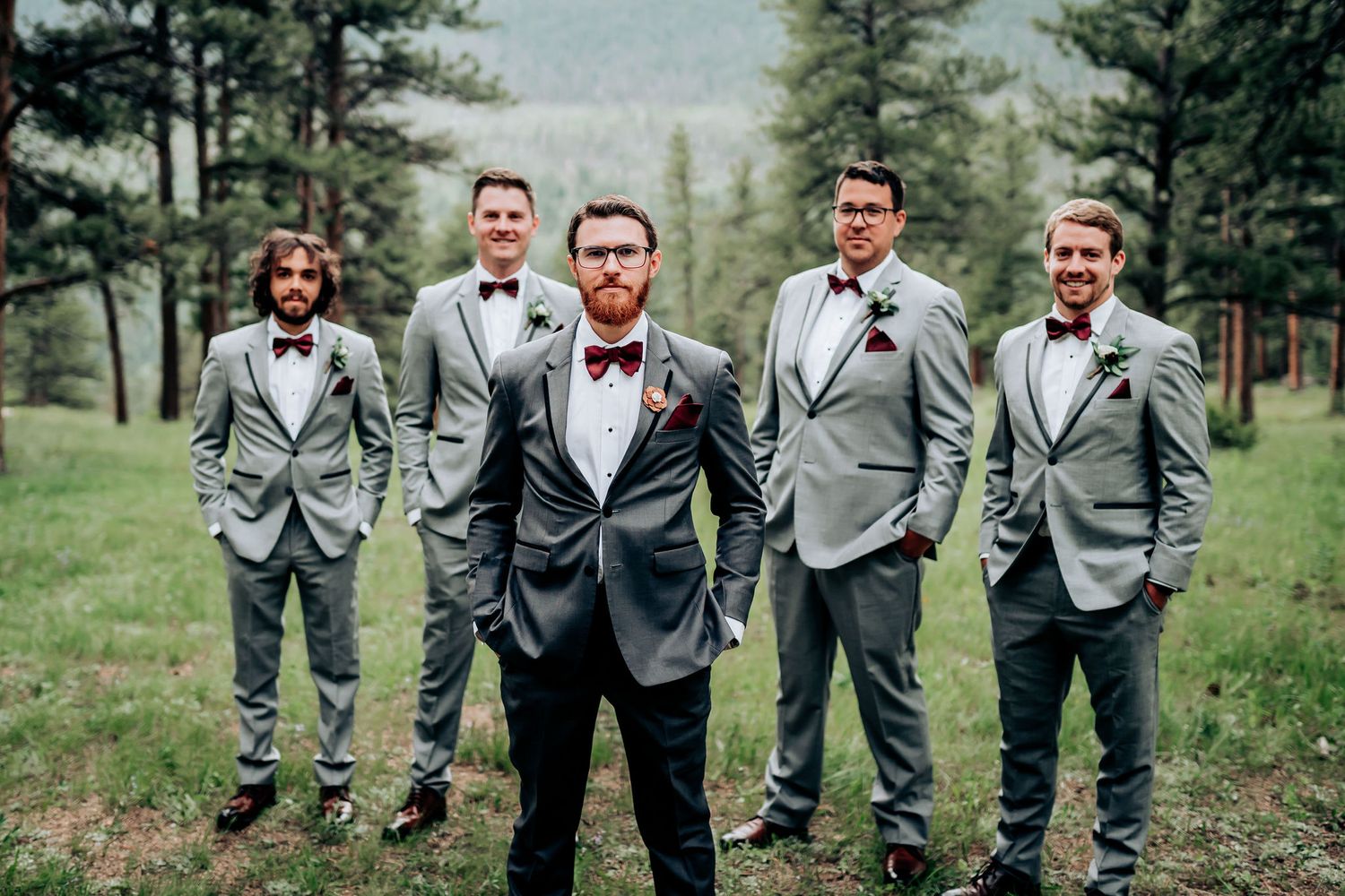 Groomsmen in gray suits and black bow ties pose in mountain forest setting.