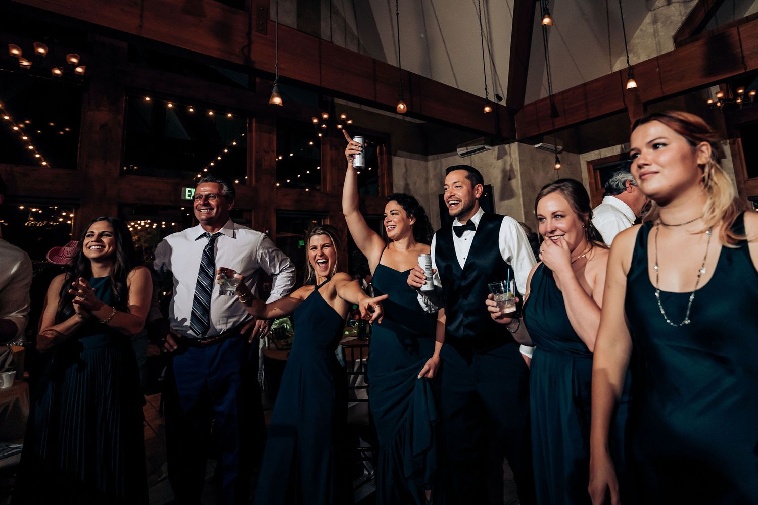 Party guests in formal attire dancing and celebrating at indoor venue with string lights.