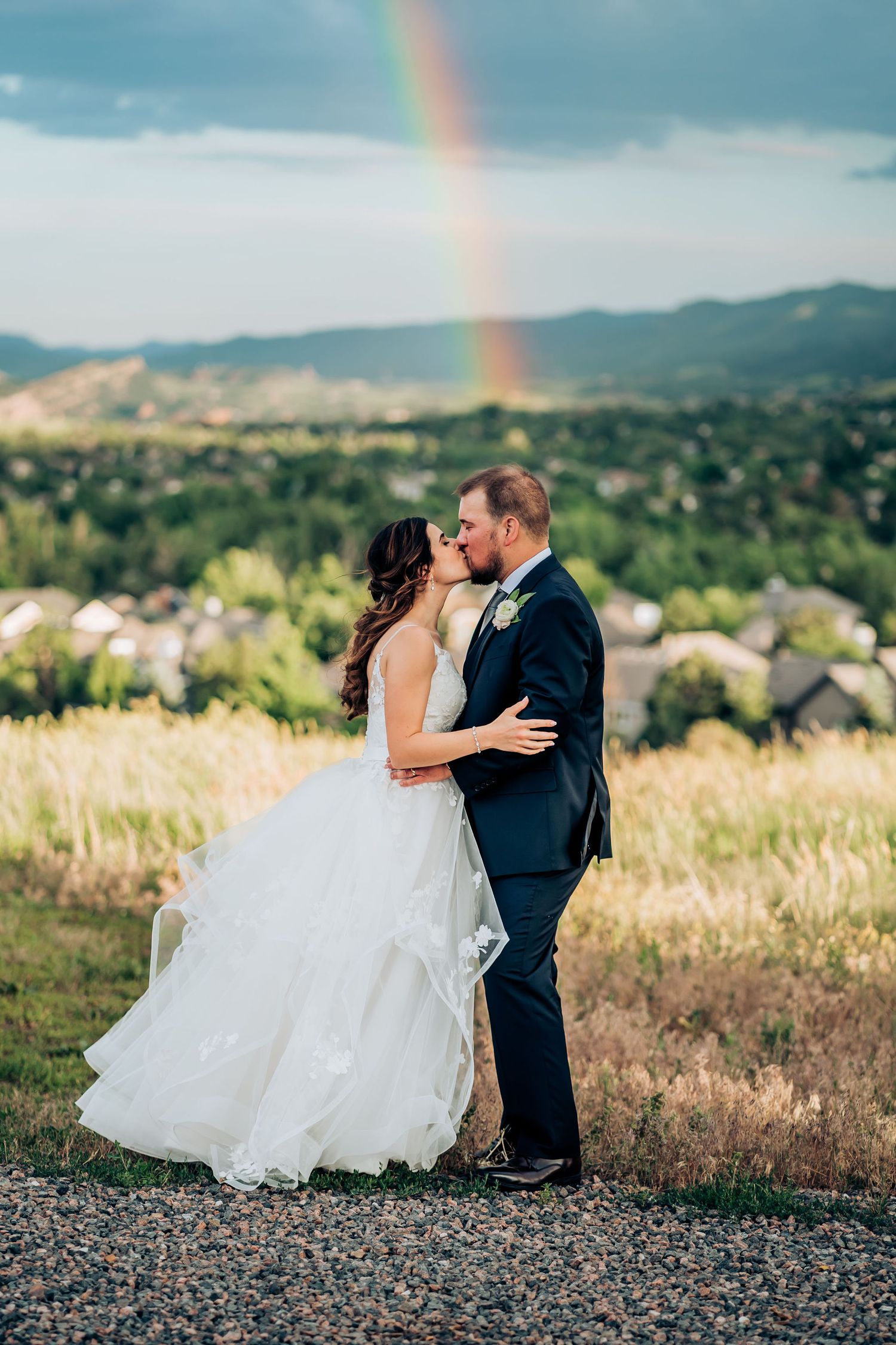 Wedding couple embraces on hilltop with dramatic rainbow and mountain landscape in background.