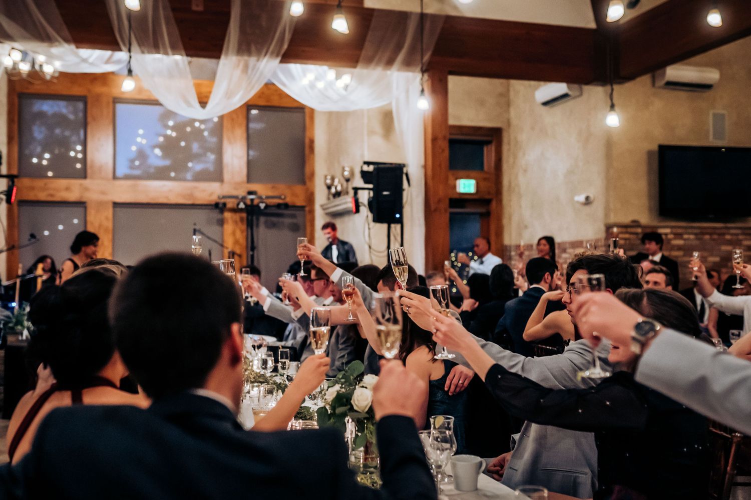 Wedding guests raising glasses for a toast in rustic venue with hanging lights.