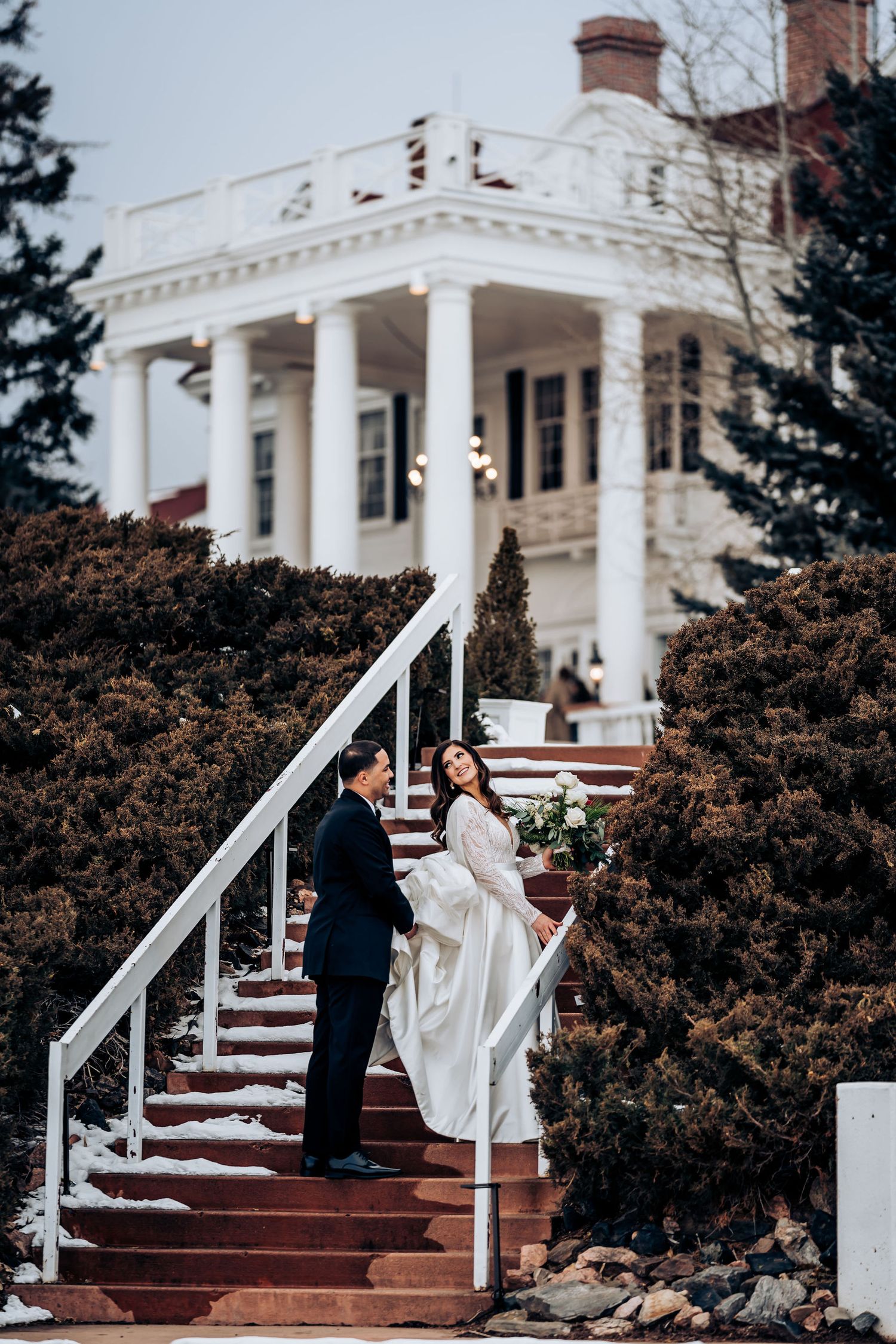 A couple stands on white mansion steps with grand white pillars on their wedding day.