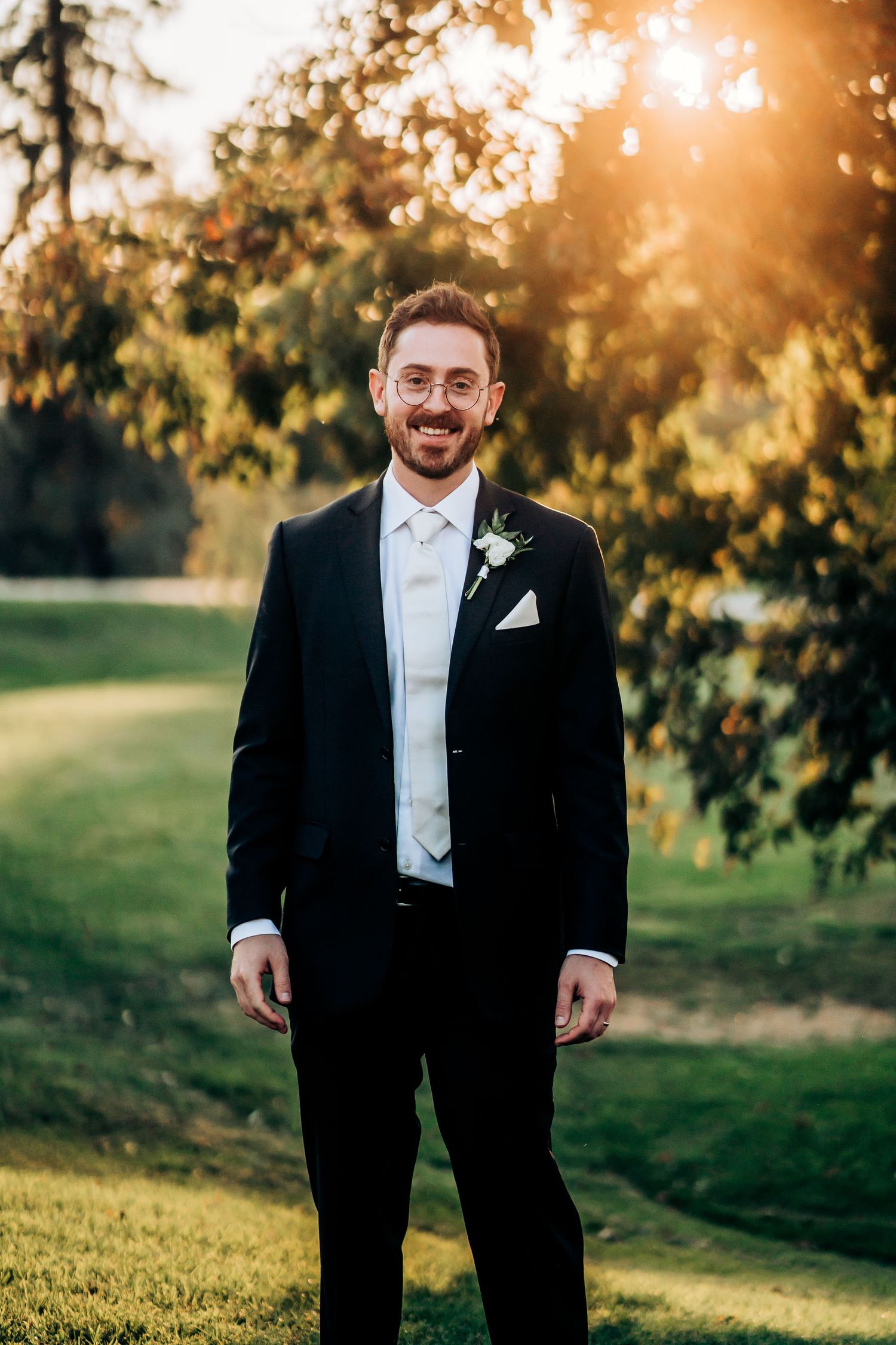 Groom in formal black tuxedo smiling warmly during outdoor sunset photoshoot.