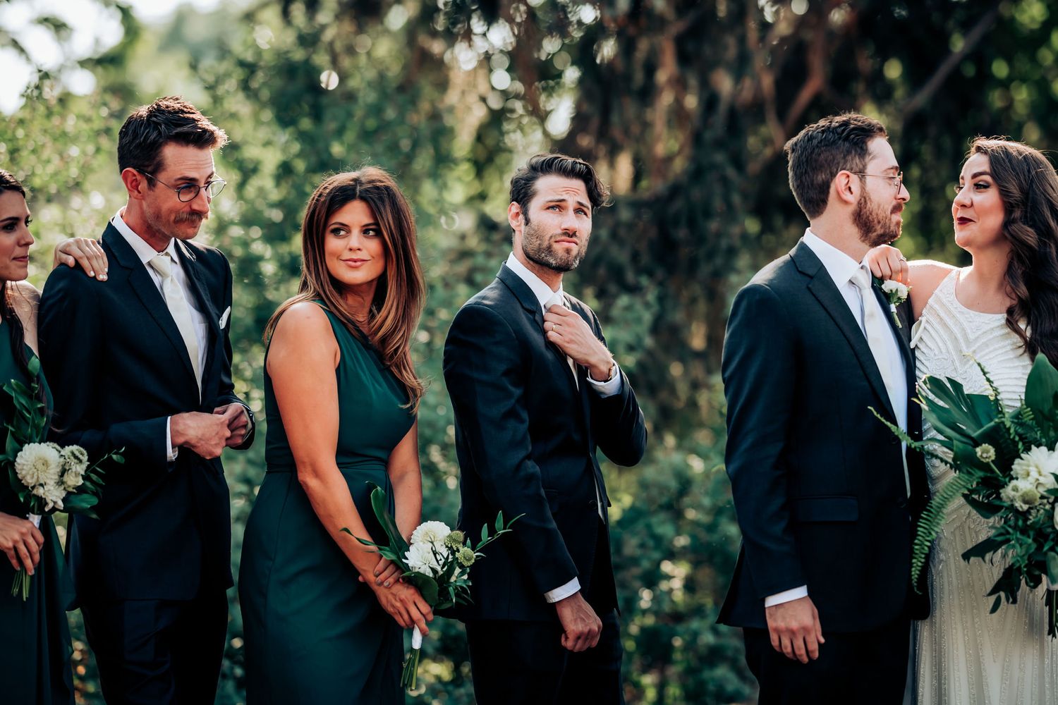 Wedding party members in dark suits and green dresses pose together in an outdoor garden setting.