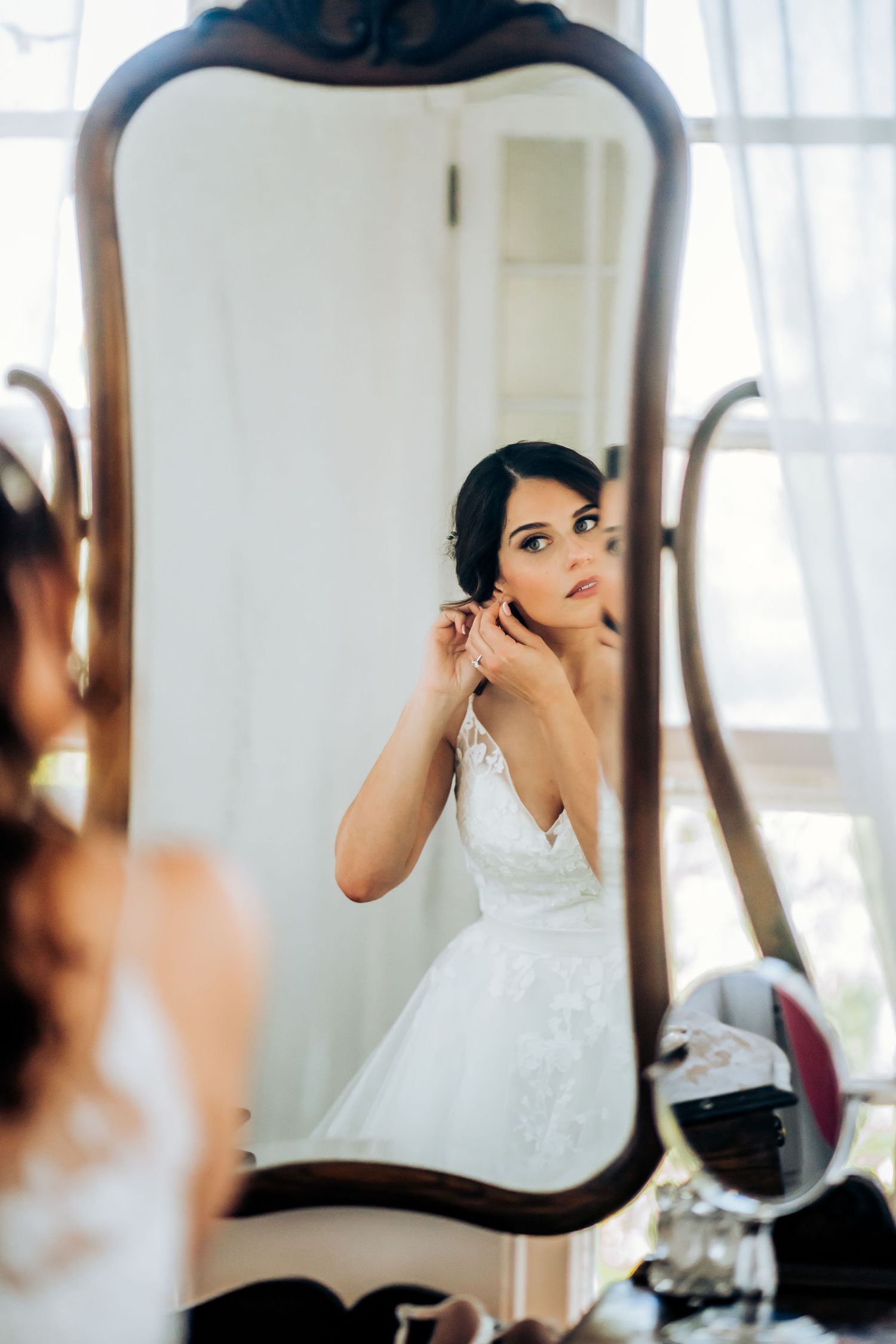 Bride in white dress puts on earrings while looking in ornate vintage mirror.