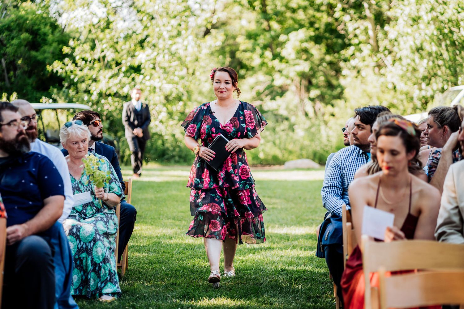 A person in a floral dress walks down an outdoor aisle between rows of seated guests.
