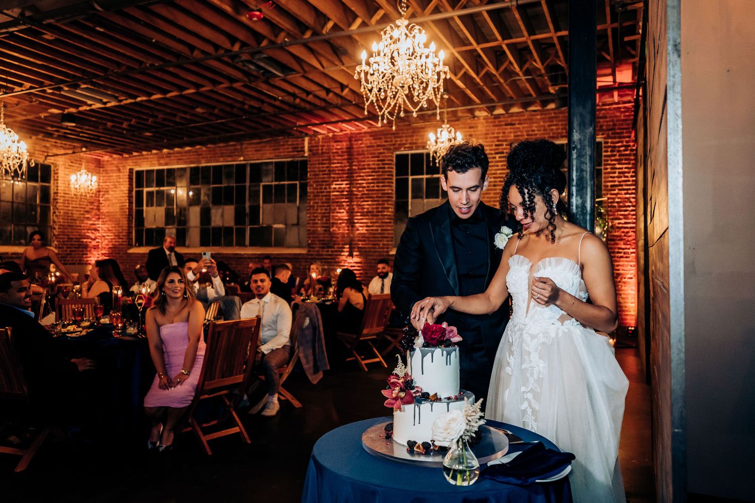 A couple cuts their wedding cake under a crystal chandelier in an industrial-style reception venue.