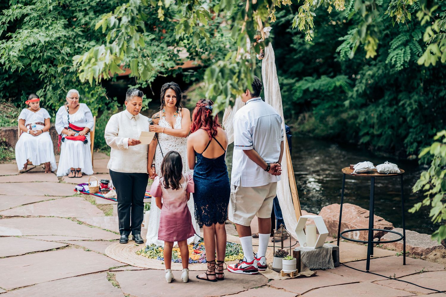 Small group gathering under wooden arch structure during outdoor wedding ceremony.