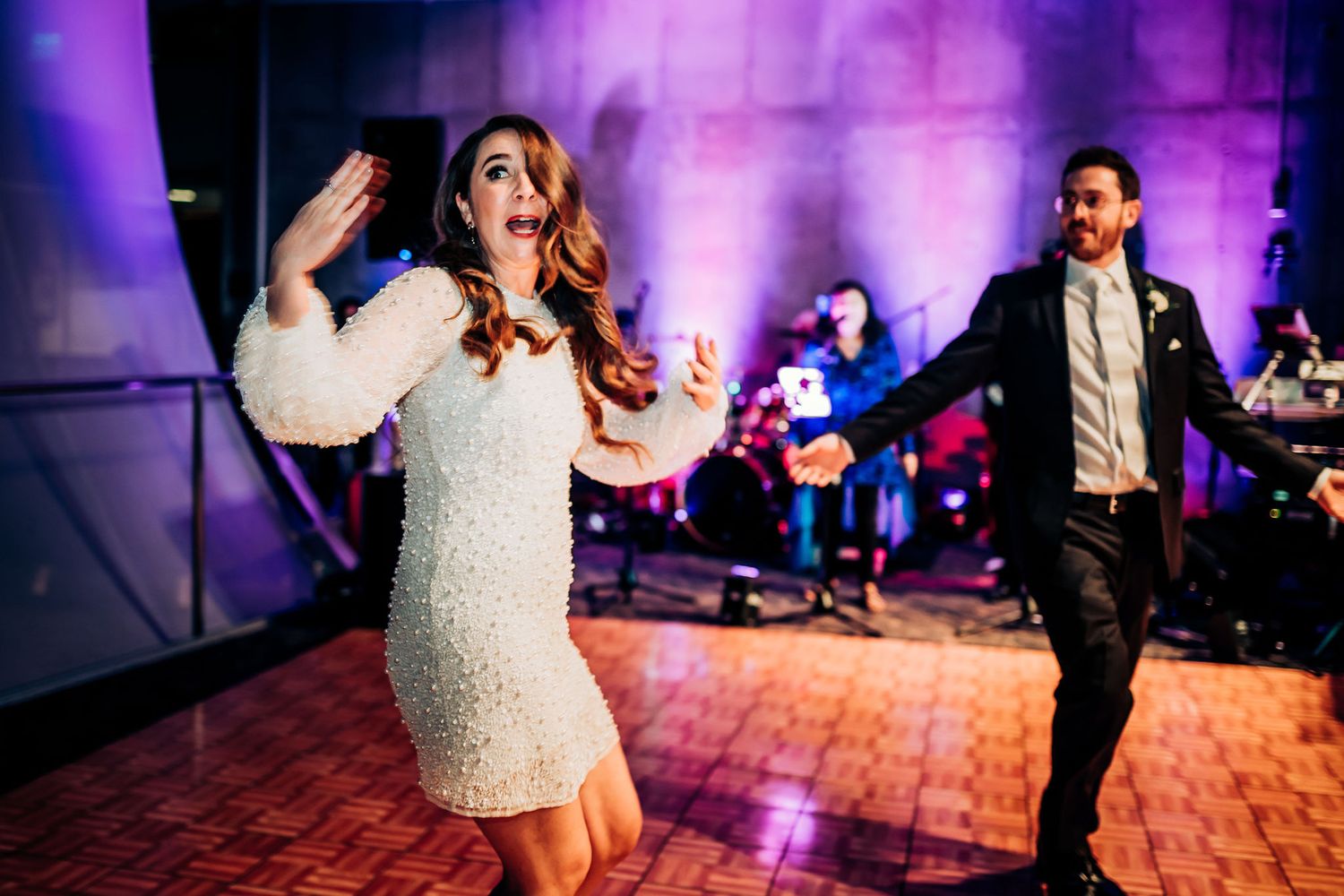 Elegant couple dancing energetically at a wedding reception with purple lighting and live band in background.