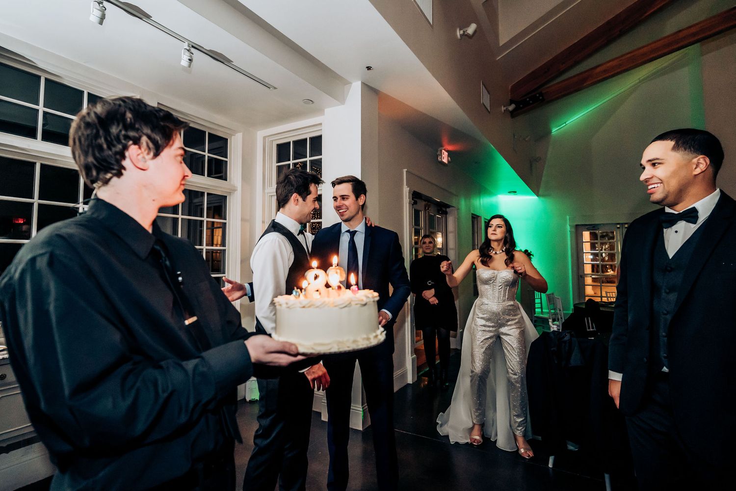 Wedding reception party with cake and guests mingling in venue with green accent lighting.