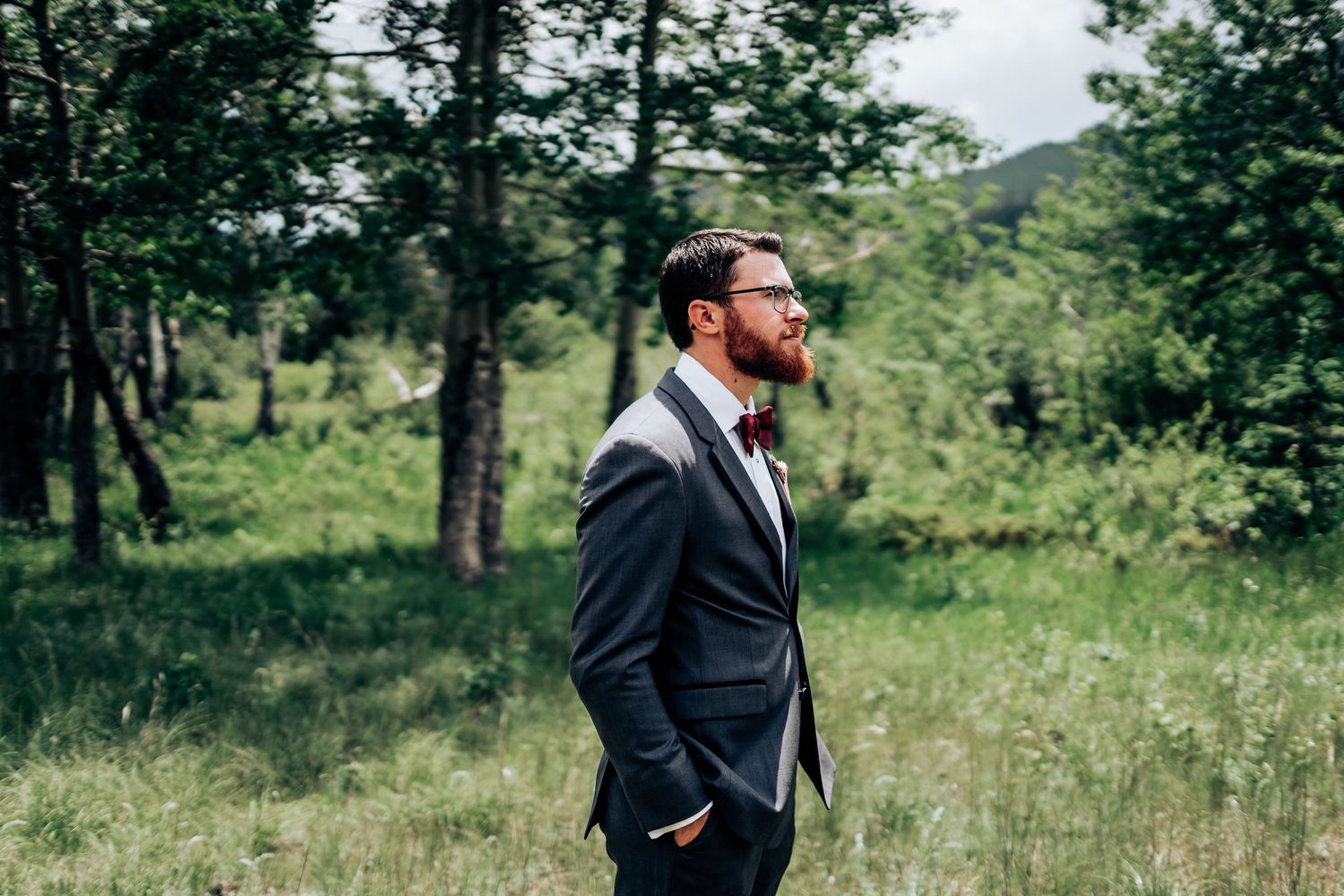 A groom in a dark suit stands thoughtfully in a scenic mountain meadow surrounded by trees.