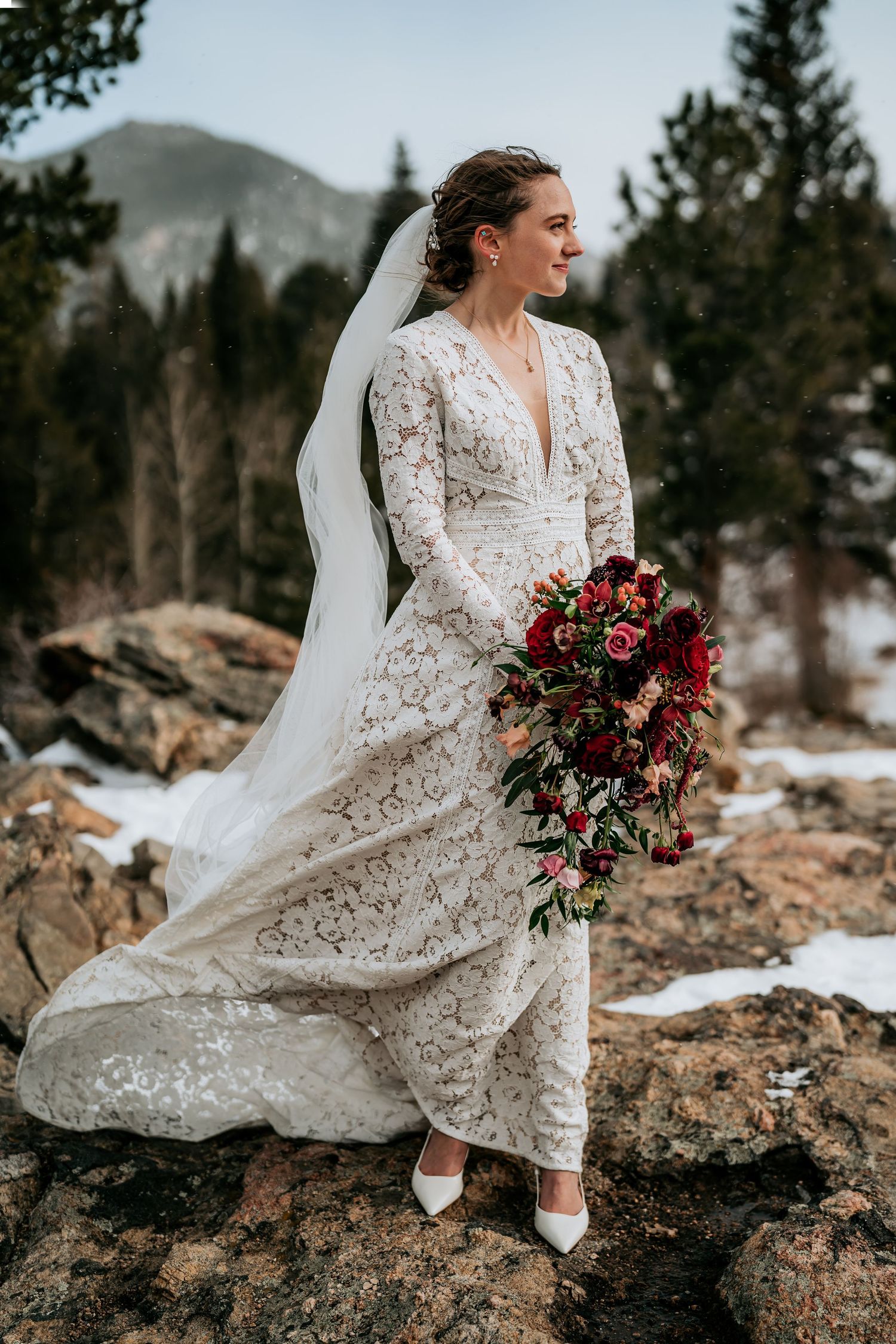 A bride in a lace wedding dress sits on rocky terrain holding a deep red bouquet.