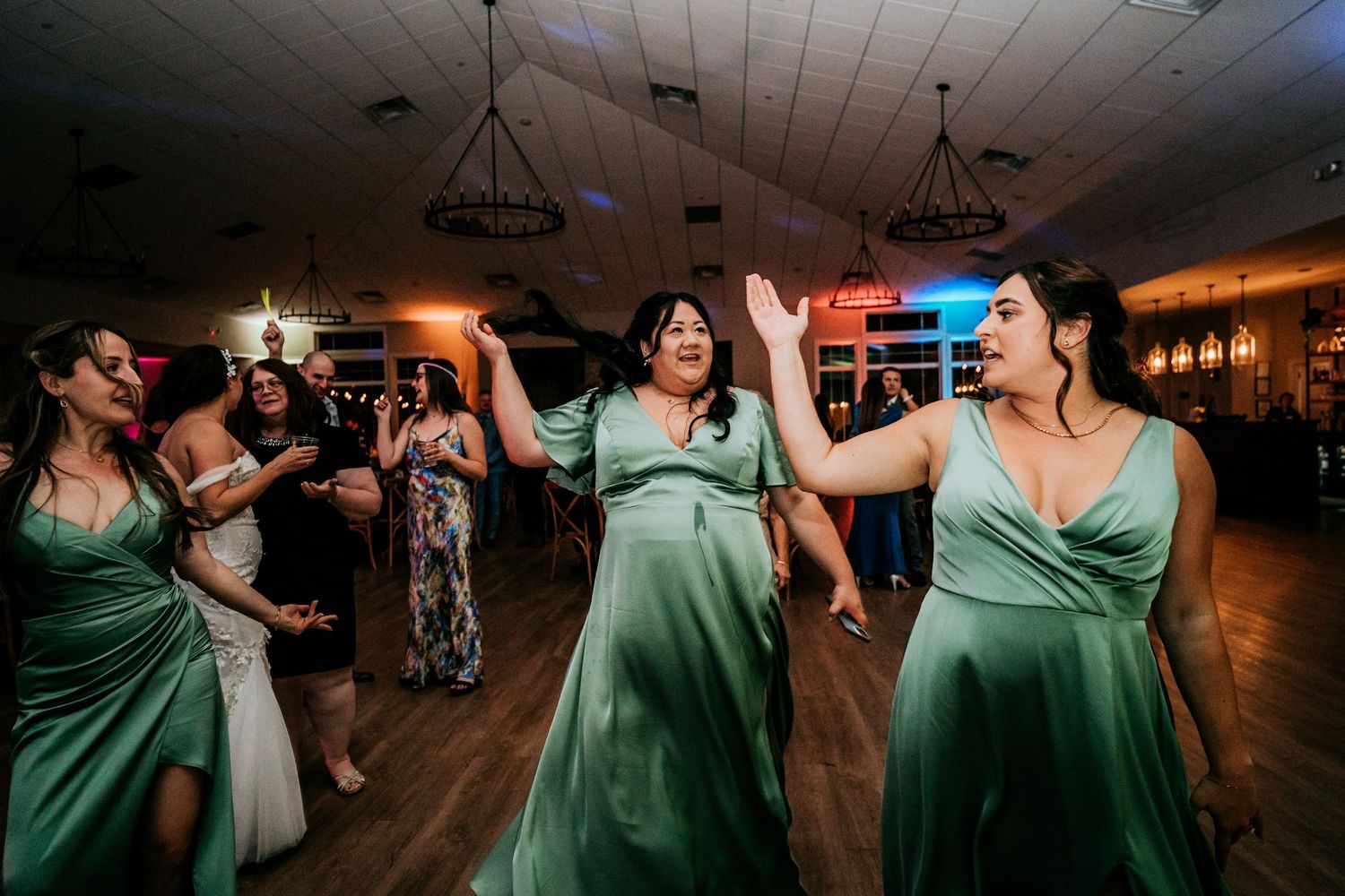 Bridesmaids in matching sage green gowns dancing together at a wedding reception.