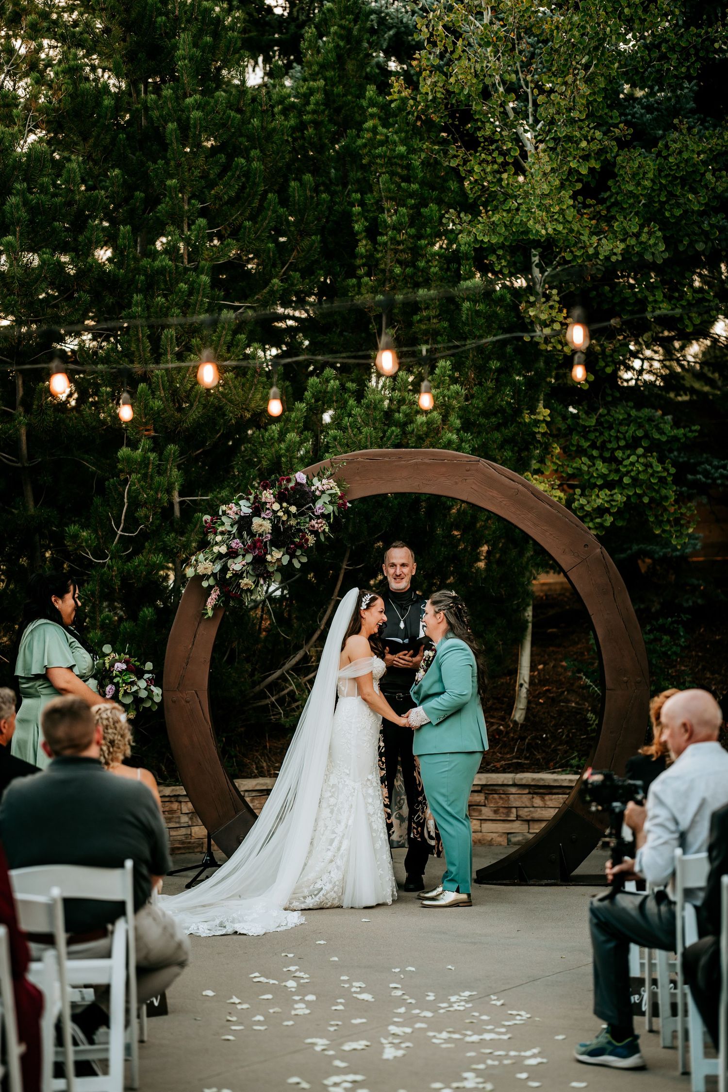 Two brides hold hands at altar under circular arch with string lights at outdoor wedding.