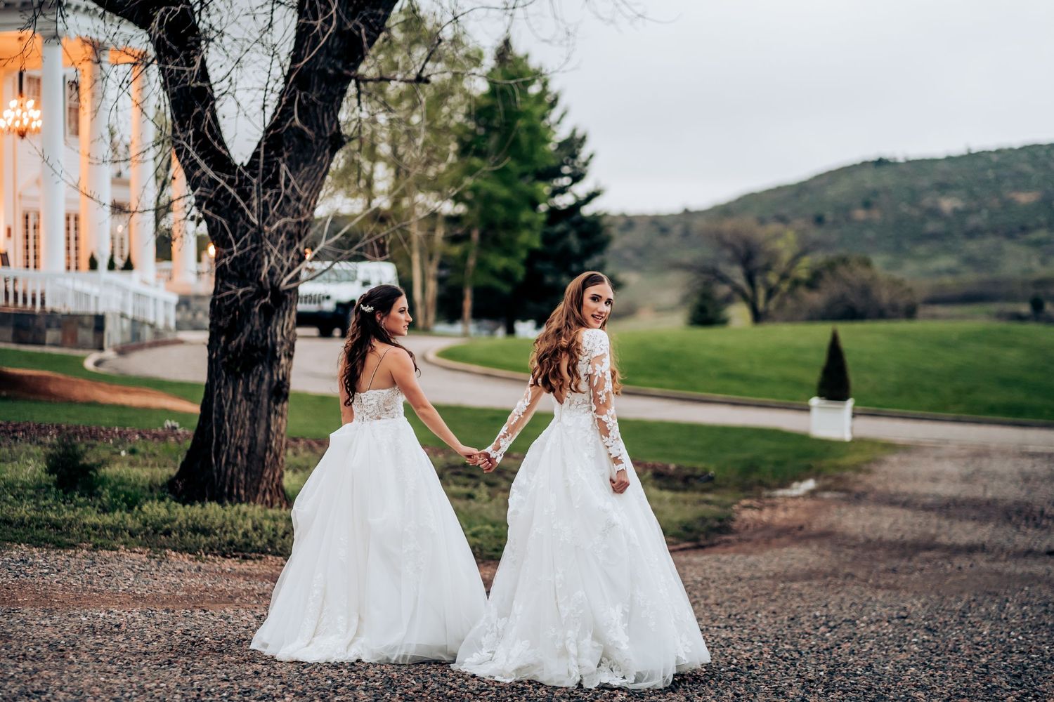 Two people in white dresses walking together on a path with mountains in background.