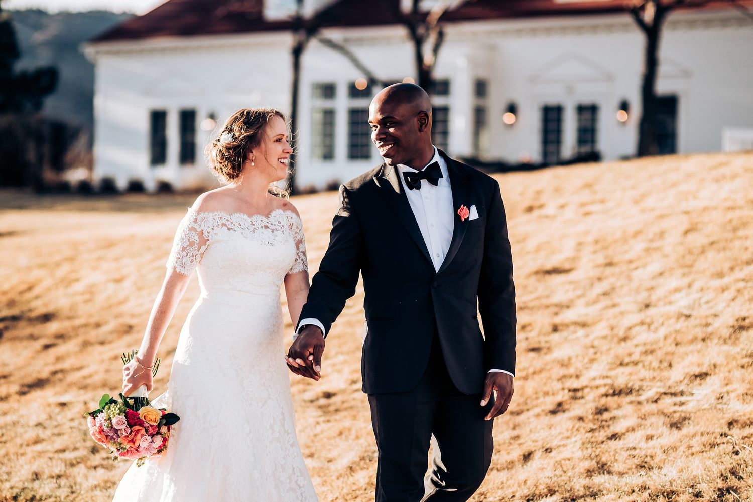 Wedding couple poses on golden hillside with white house in background.