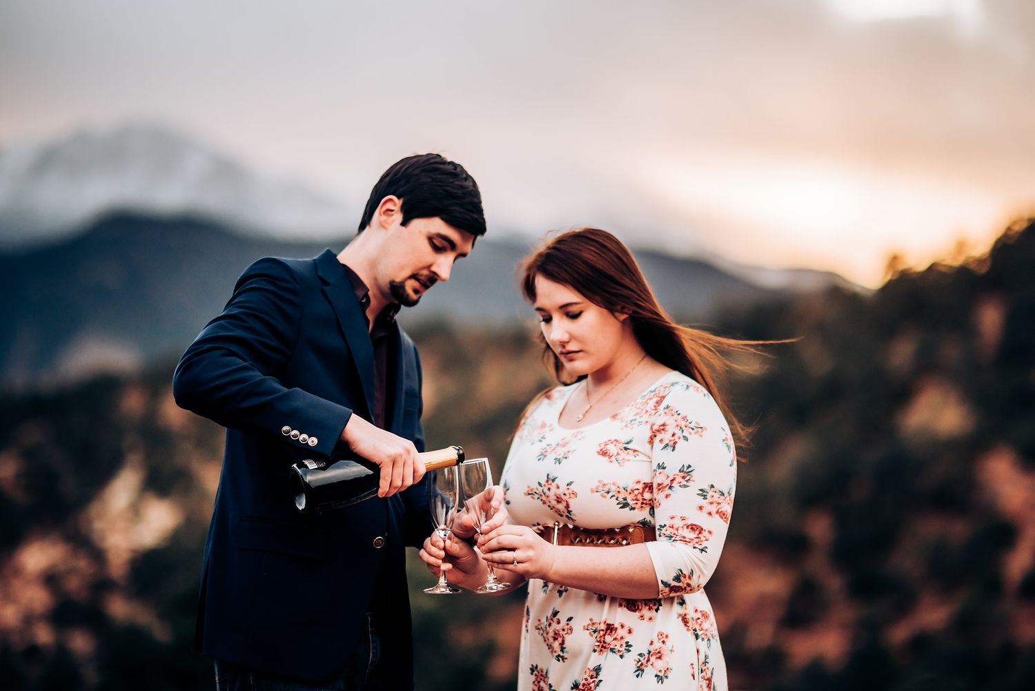 A couple opens champagne together during sunset on a mountain overlook.
