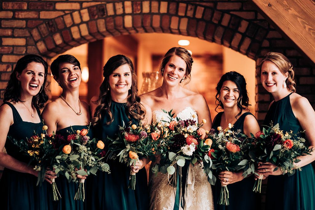 Wedding party in emerald green dresses holding lush bouquets under warm lighting.