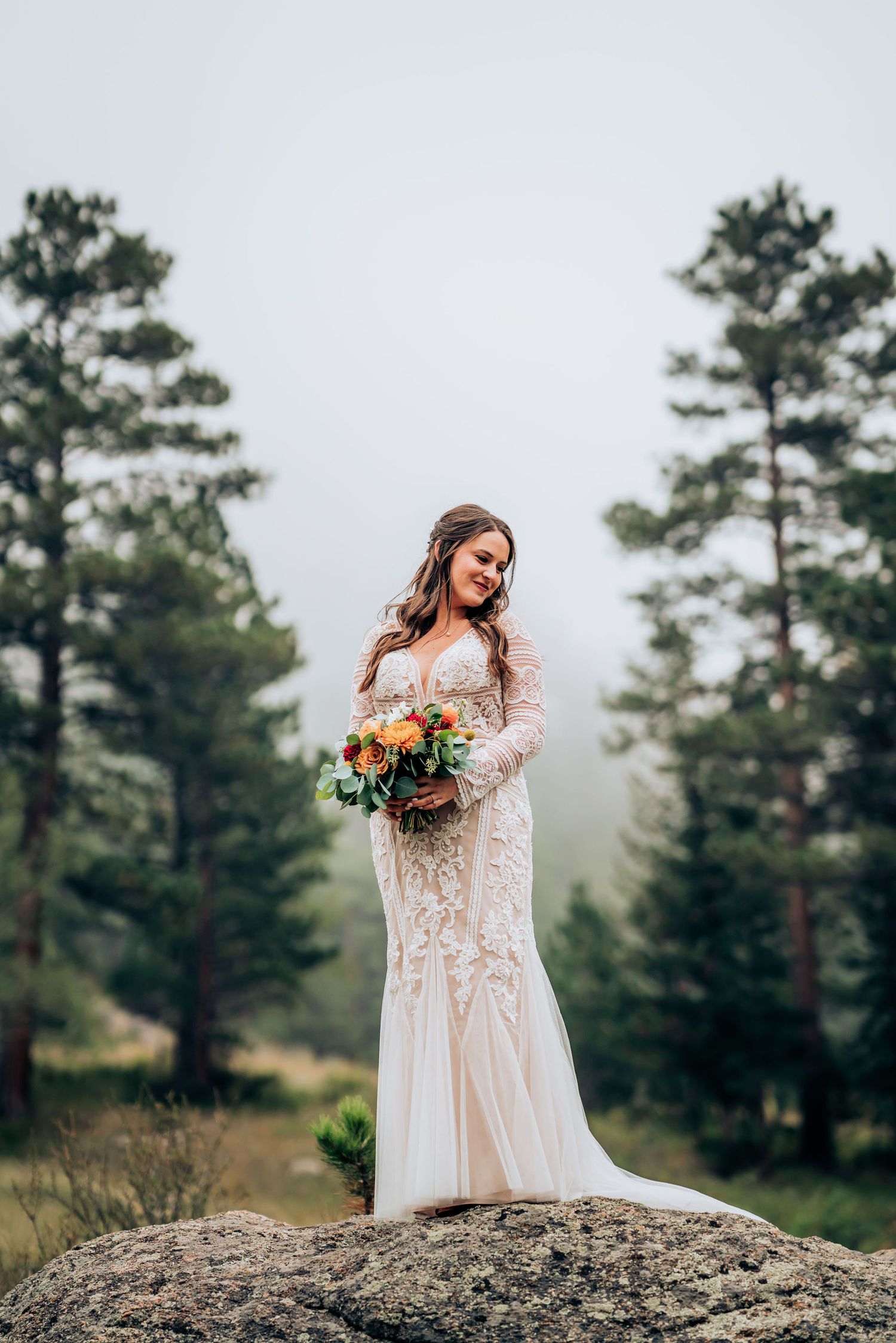 A bride in a lace gown holds a bouquet while standing on a rocky overlook.