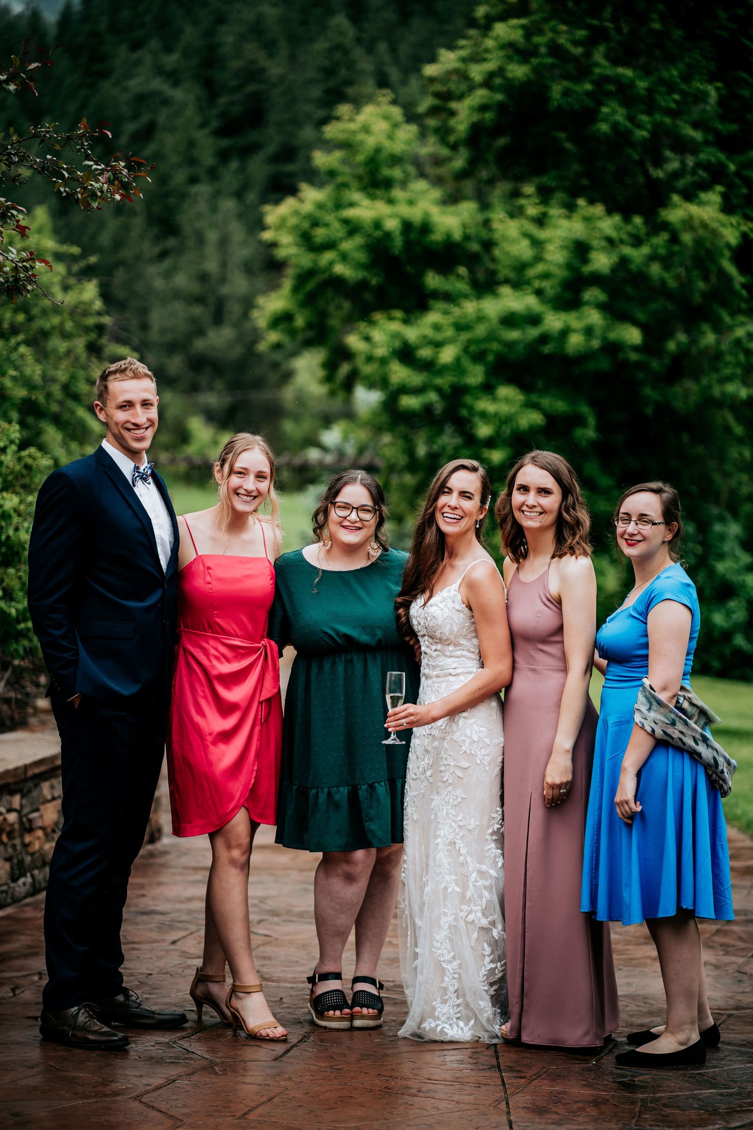A wedding party in coordinated formal attire poses on a stone pathway surrounded by green forest.