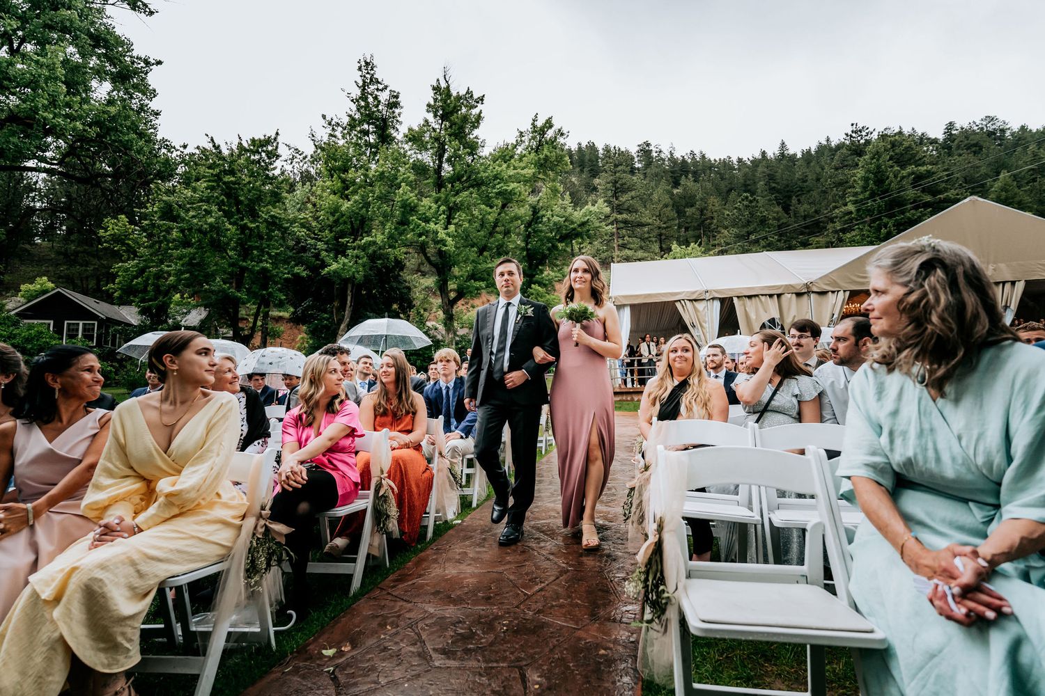 Wedding guests seated on white chairs during an outdoor ceremony with trees in the background.