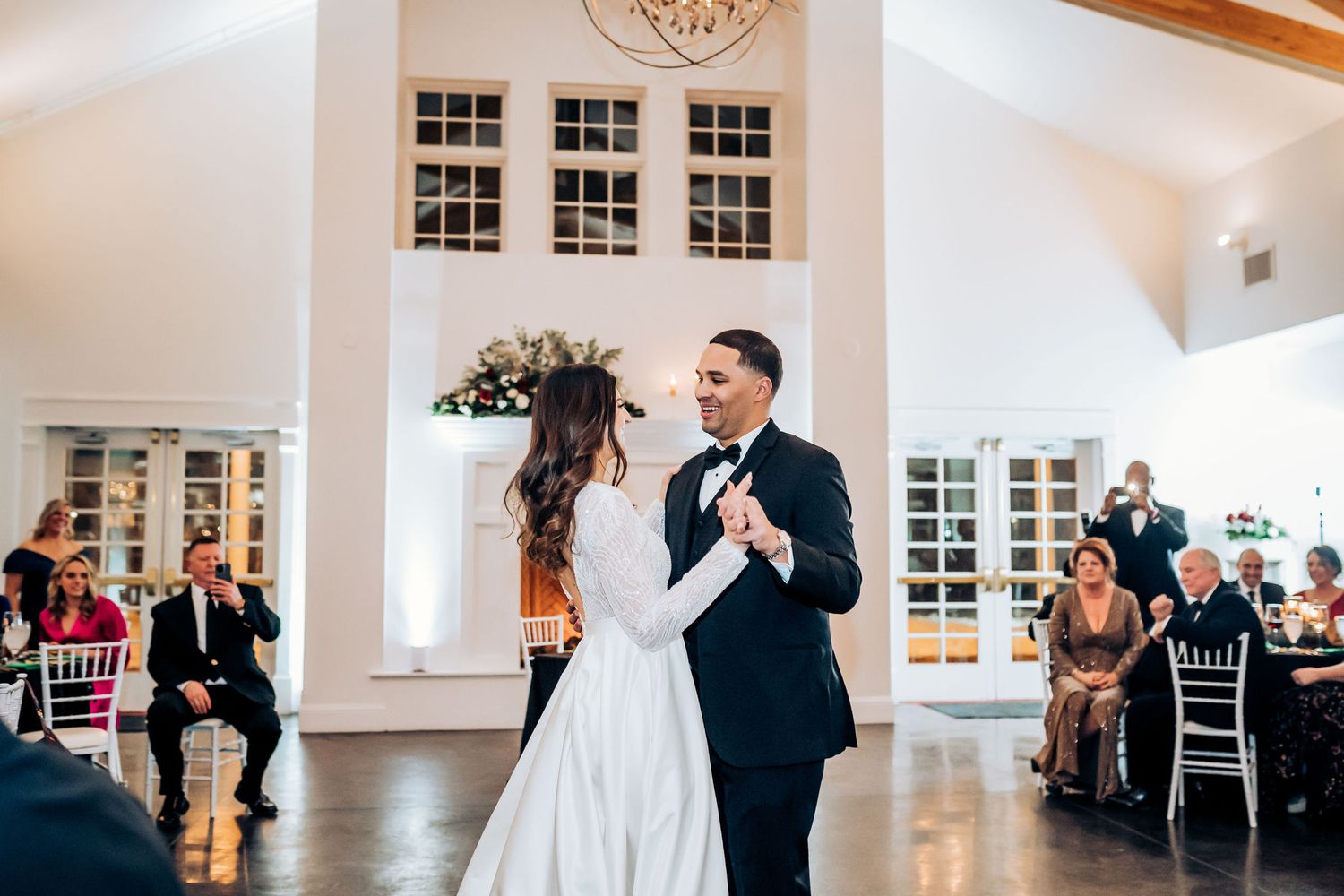 Wedding couple dances in white reception hall with high ceilings and wooden beams.