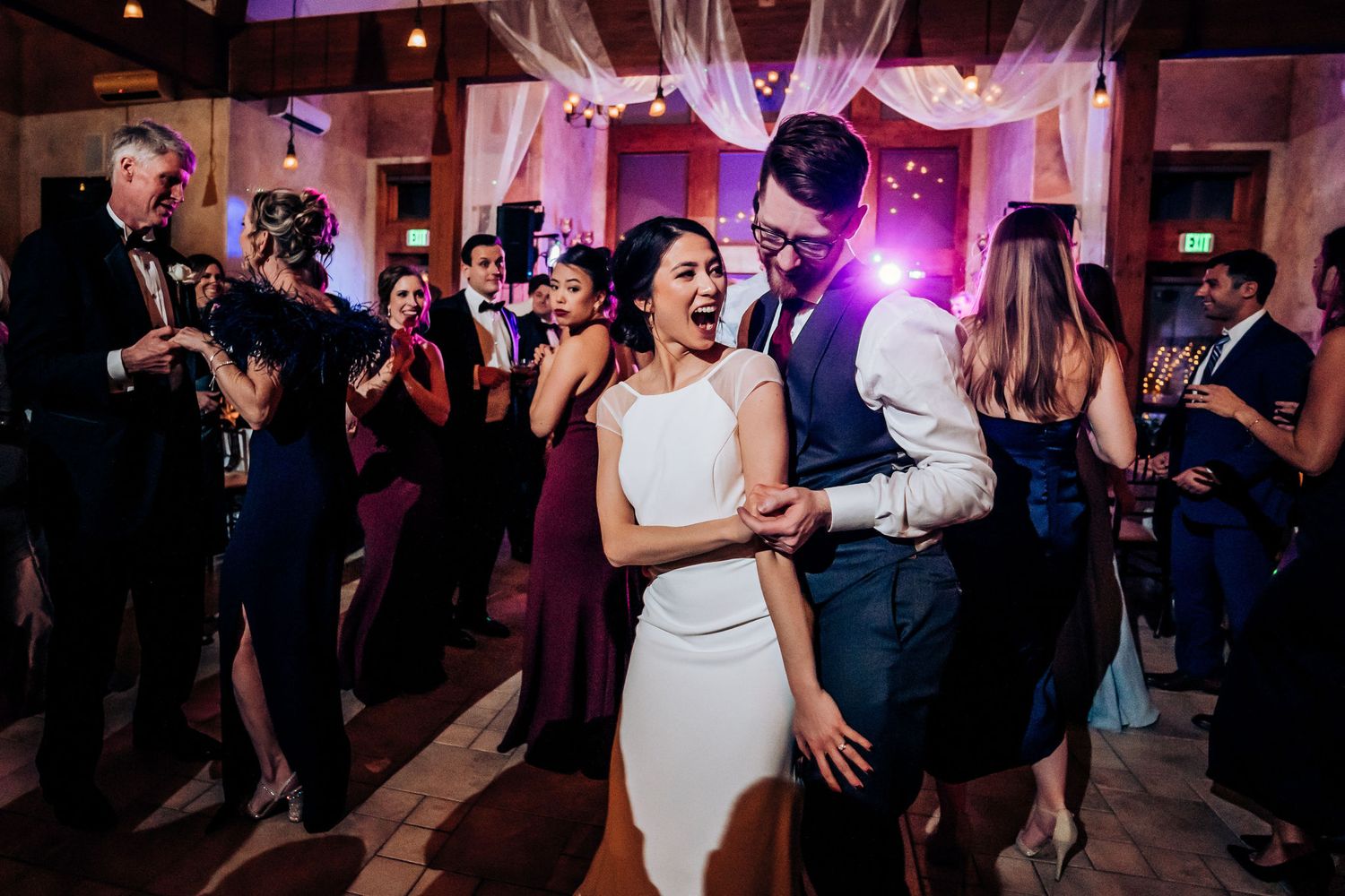 A couple dances together on their wedding reception dance floor surrounded by guests and pink mood lighting.