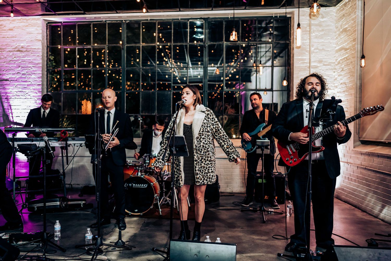 Full wedding band performs on stage with string lights and modern decor.