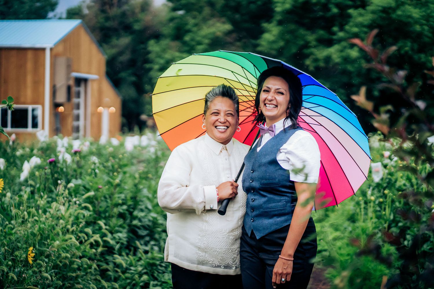 Two people pose joyfully with a rainbow umbrella in a sunny garden setting.