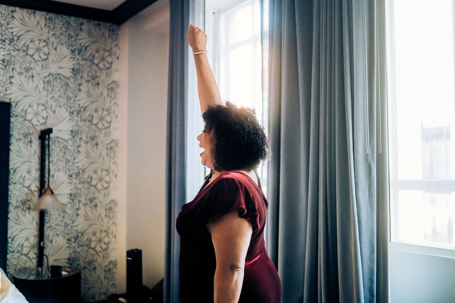 A person in burgundy reaches up to adjust curtains in a hotel room with floral wallpaper.