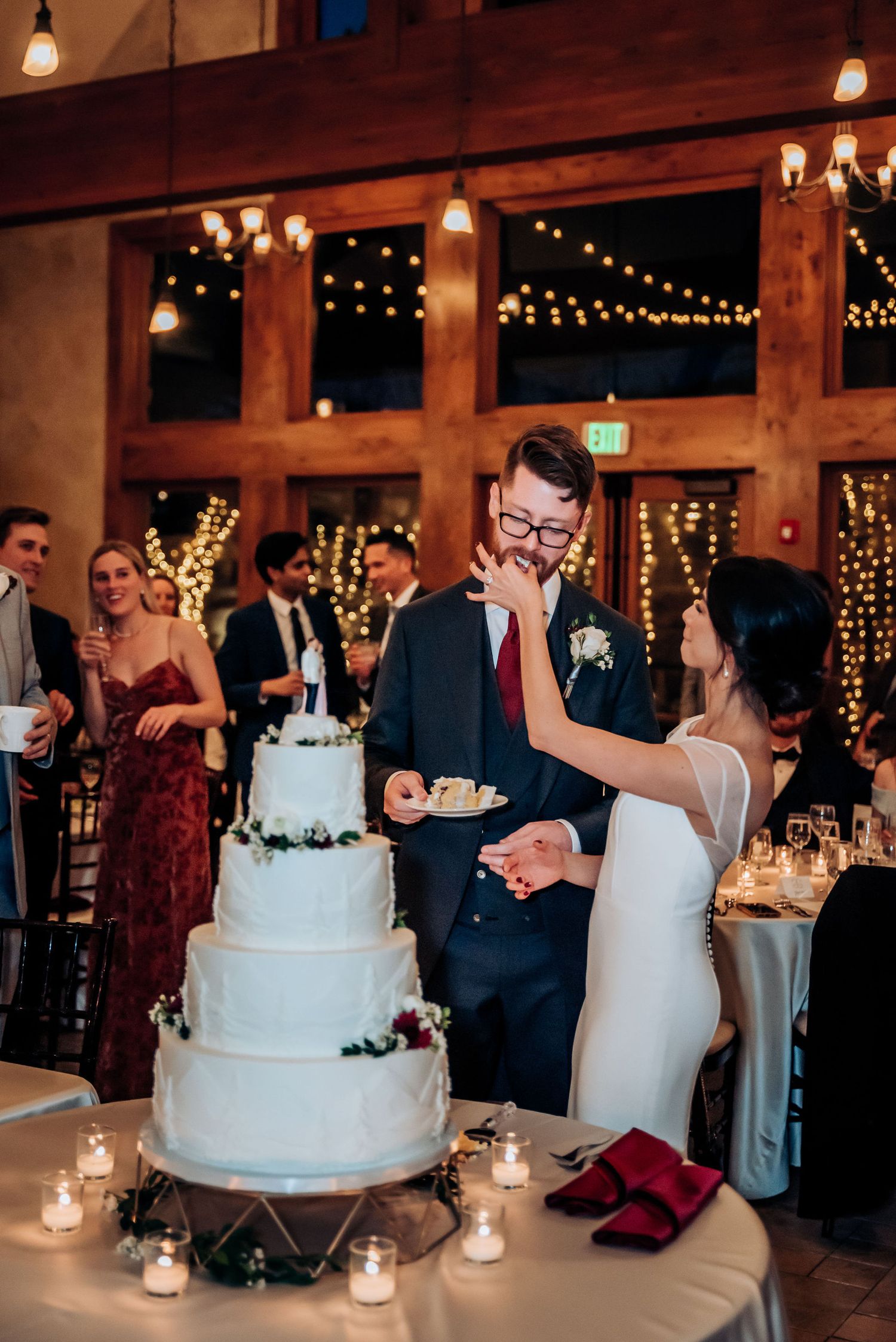 Newlyweds share cake beside their elegant white tiered wedding cake decorated with greenery.