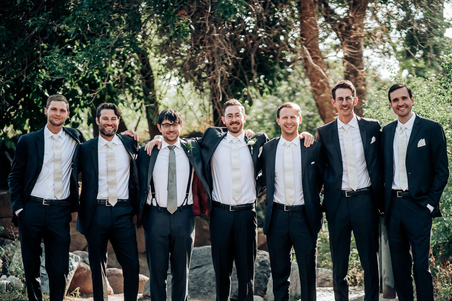 Groomsmen in black tuxedos and bow ties pose together outdoors under trees.