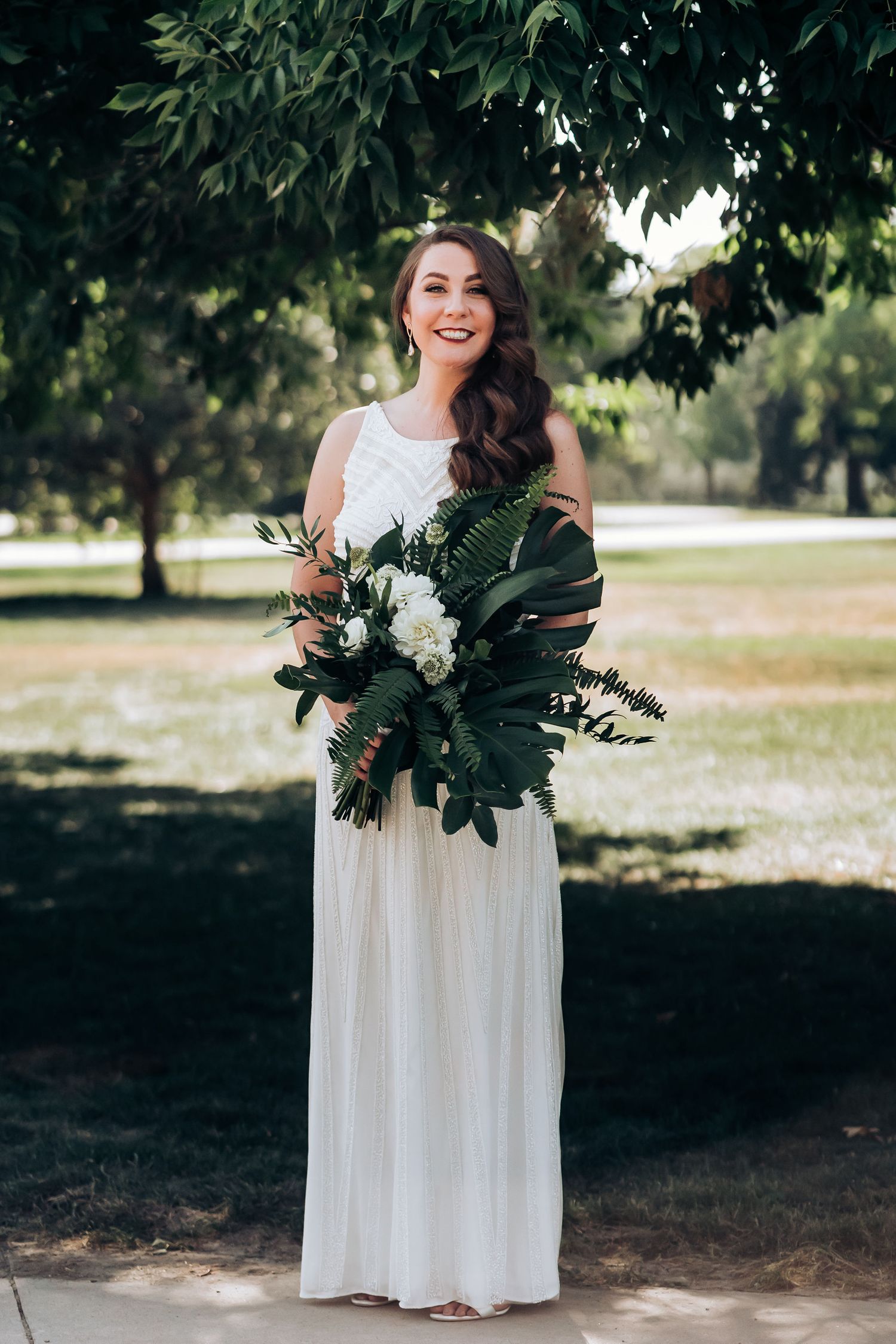 A person in a white wedding dress holds a stunning bouquet of white flowers and tropical greenery outdoors.