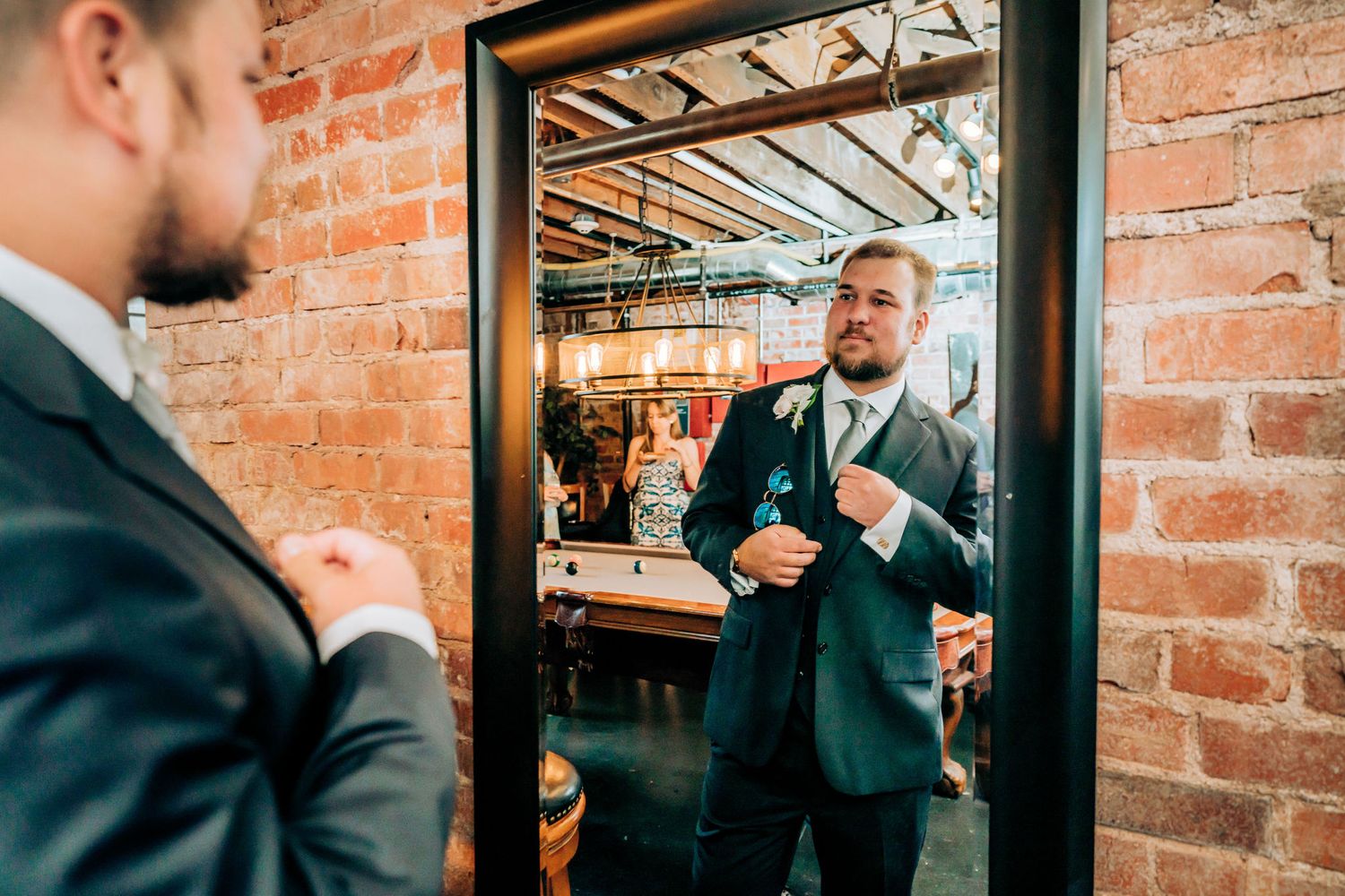 Person adjusts tie while looking in mirror with exposed brick wall background.