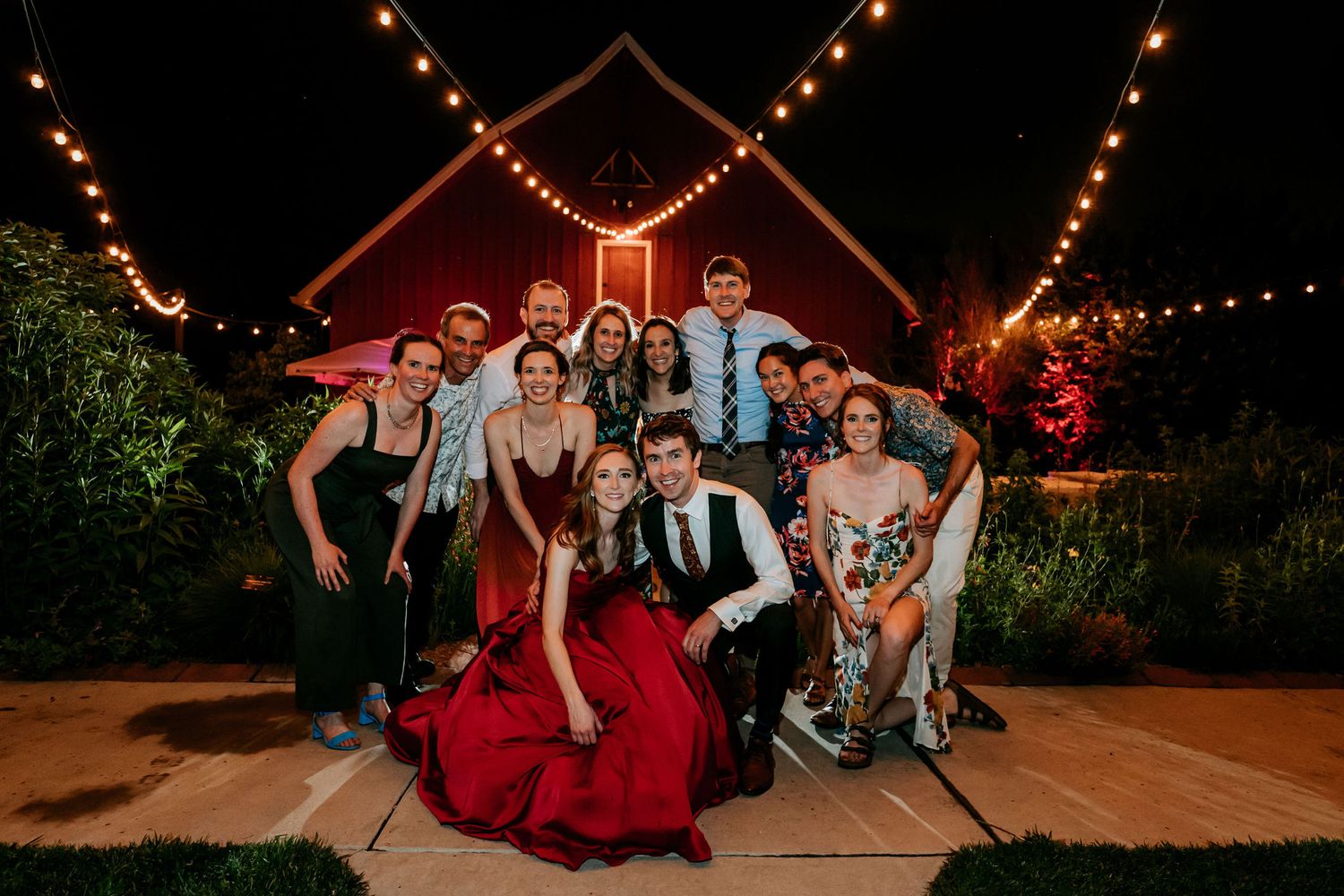 Wedding party poses at night under string lights in front of red barn.