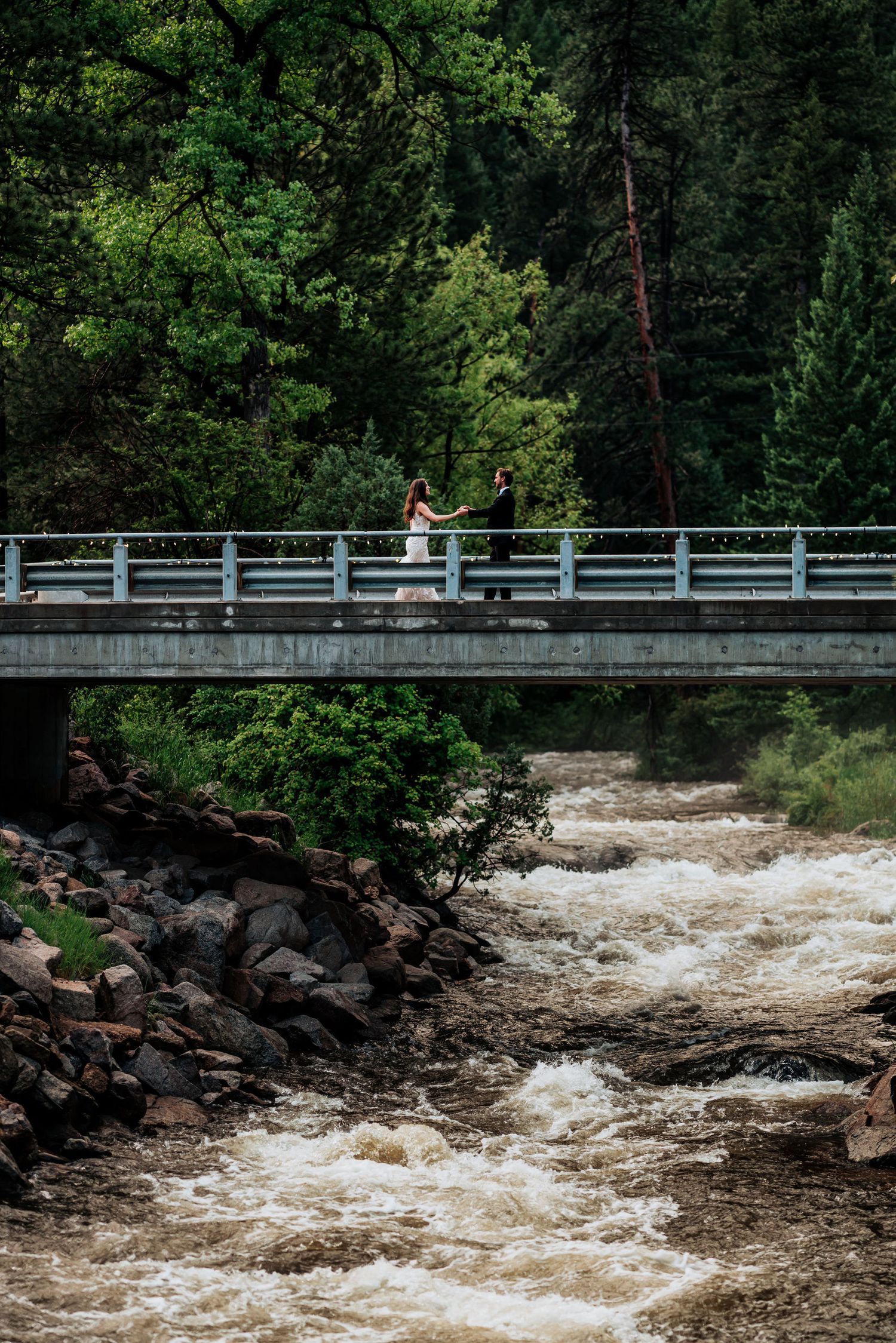 Two figures stand on a bridge over rushing rapids surrounded by lush forest.