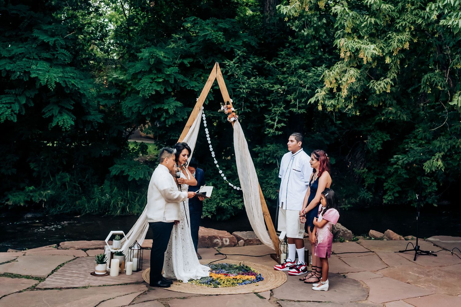 Outdoor wedding ceremony under a wooden triangle arch surrounded by lush greenery.