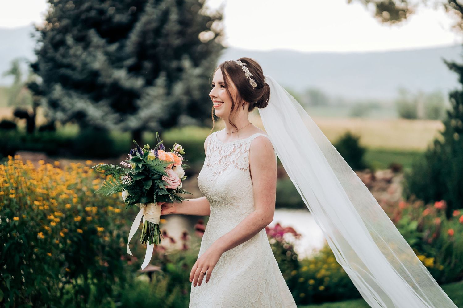 Bride in elegant lace gown with flowing veil stands among wildflowers.