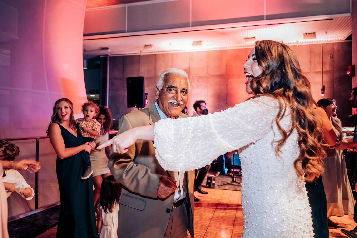 White dress dancing with guest during wedding reception under warm pink lighting.