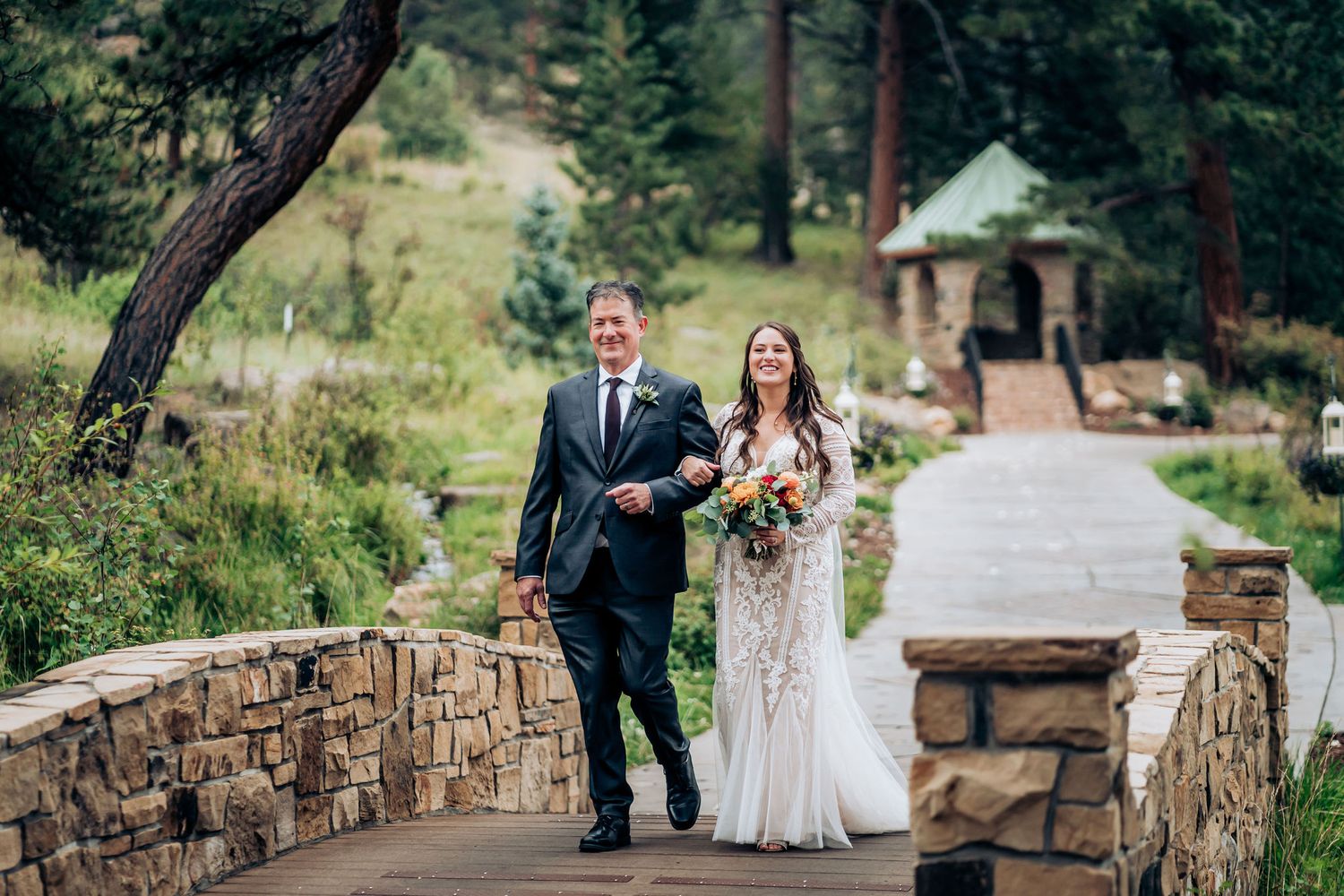 A couple walking across a stone bridge during their outdoor mountain wedding ceremony.