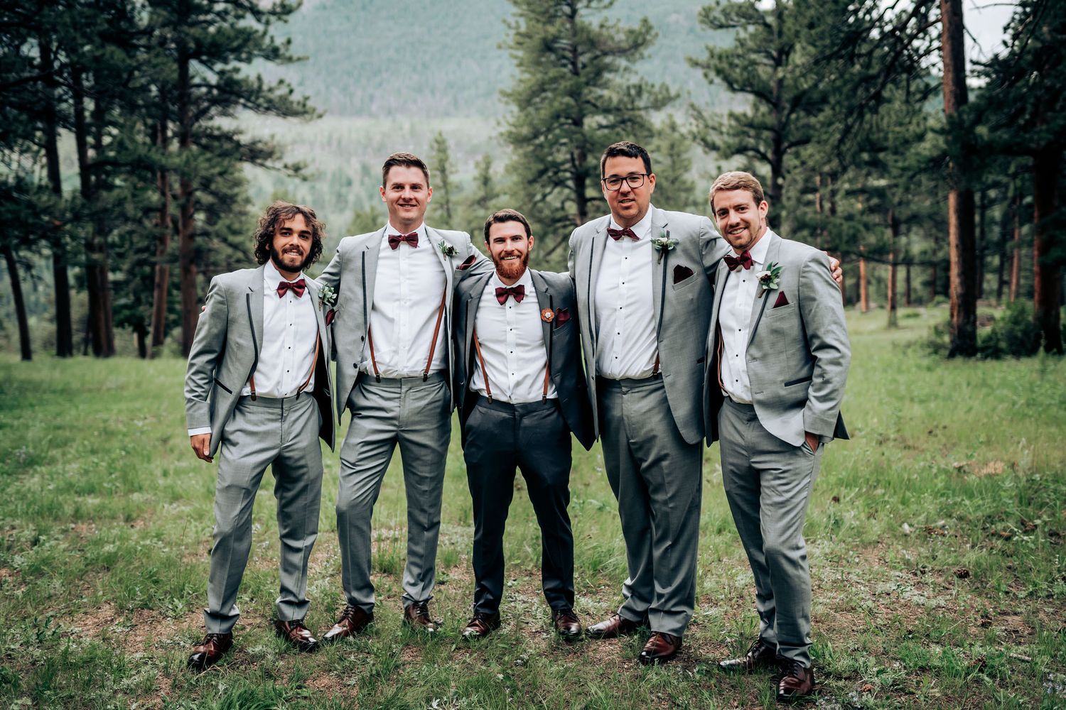 Groomsmen in gray suits and suspenders pose together in mountain forest setting.