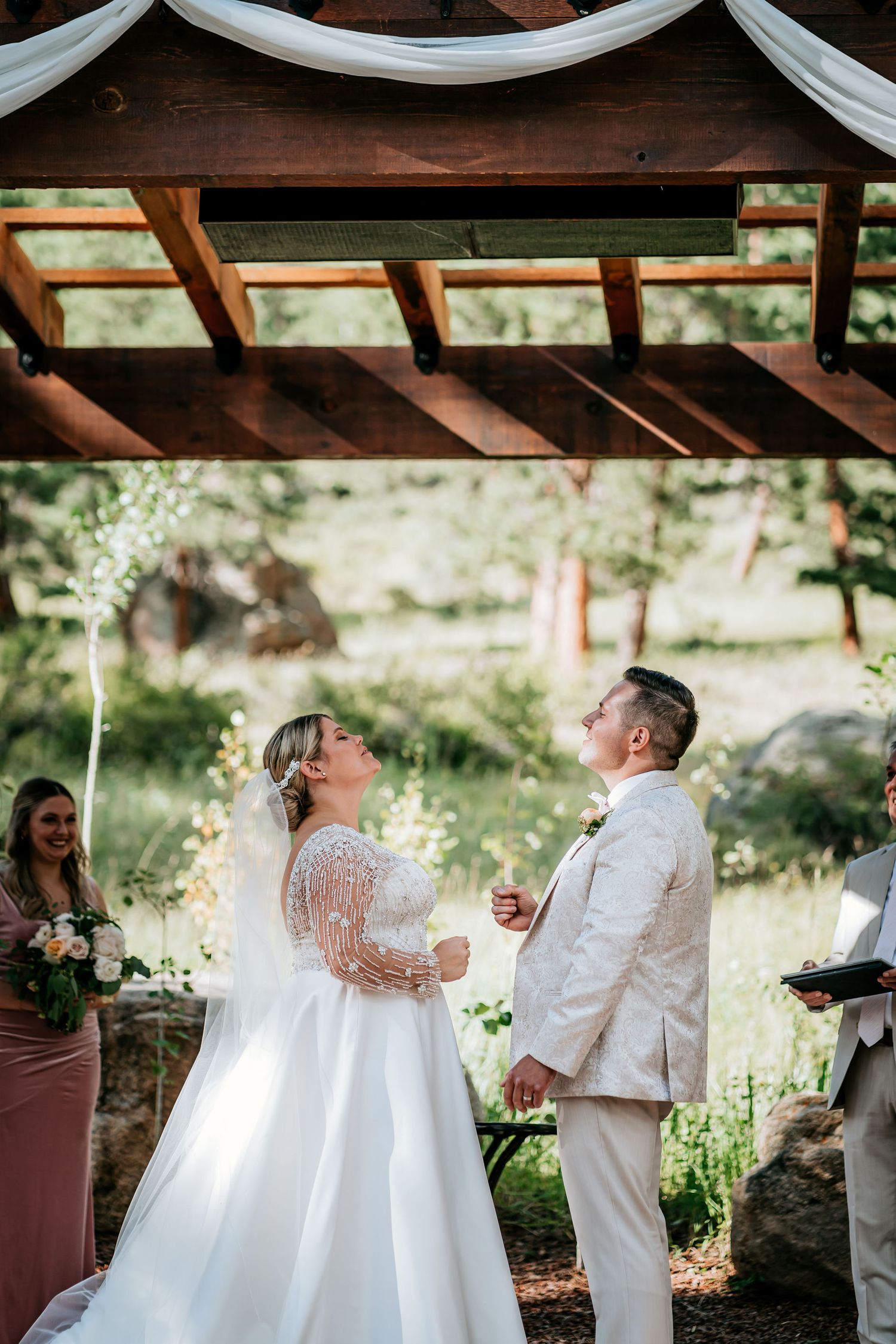 Wedding ceremony under wooden pergola with white draping and natural surroundings.