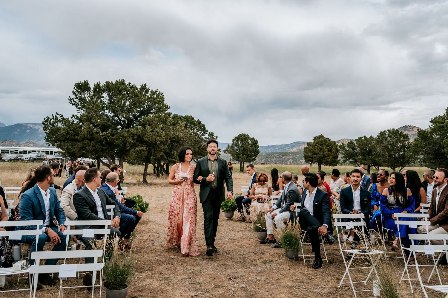 Wedding processional through rustic mountain venue with guests seated on white chairs.