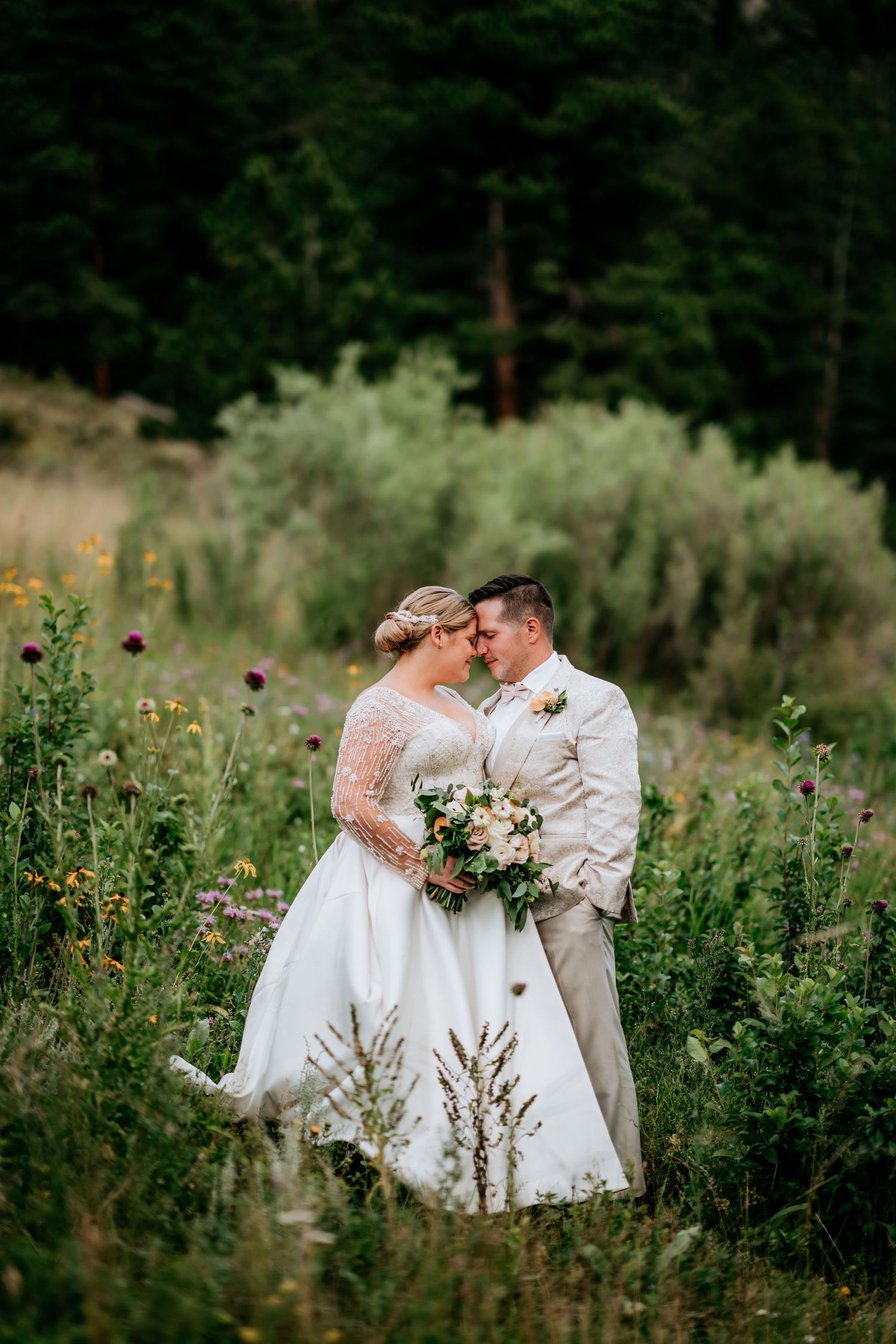 A romantic wedding couple embraces in a wildflower meadow against a backdrop of evergreen trees.