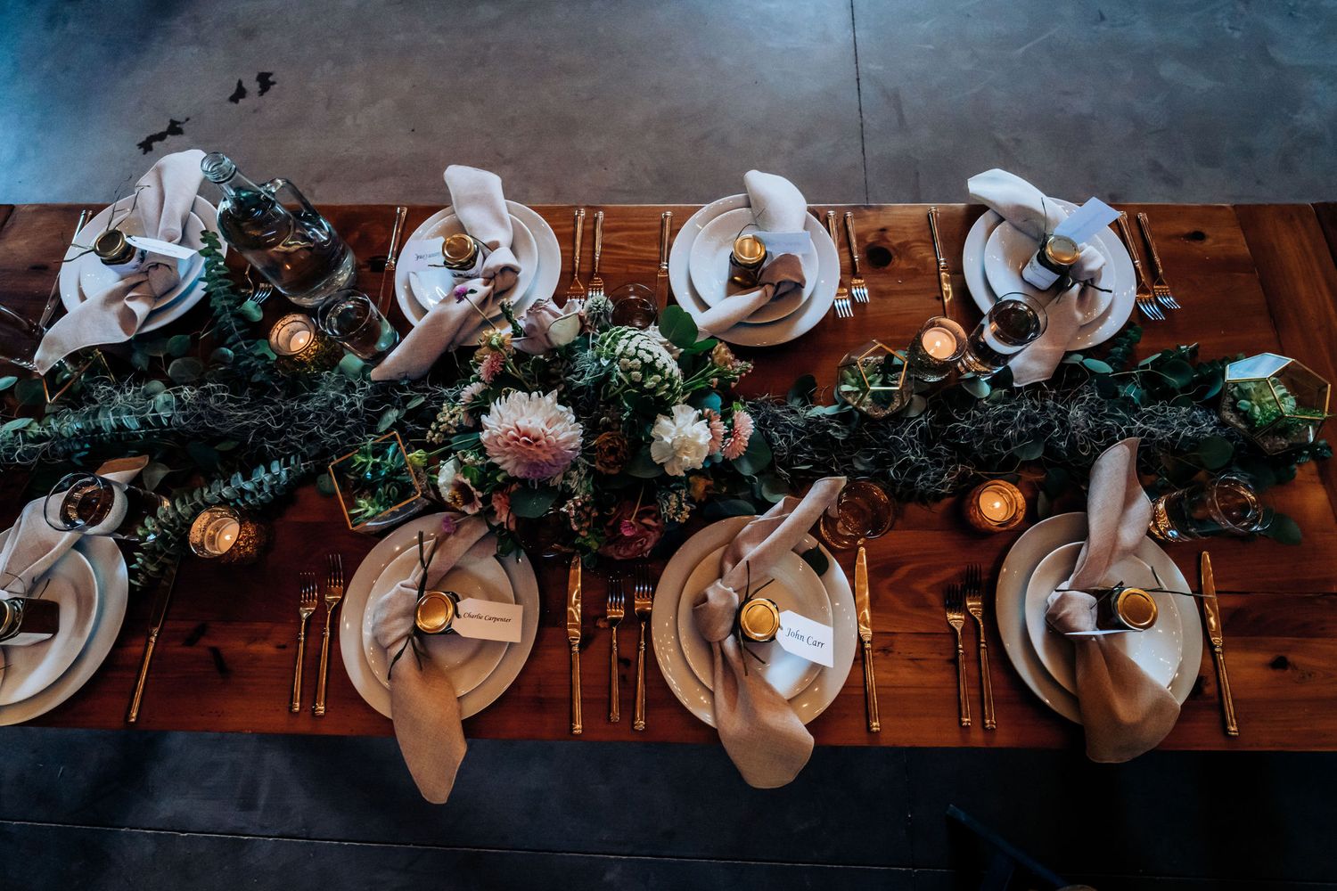 Elegant wooden table setting with floral garland centerpiece and white place settings for wedding reception.