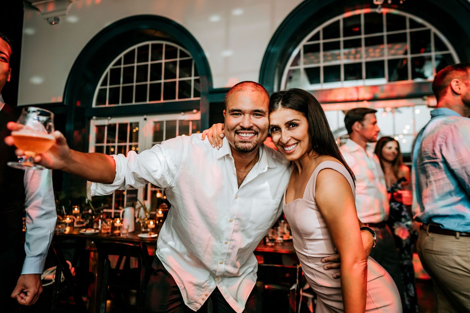 Party guests celebrating at a reception venue with arched windows and bar in background.